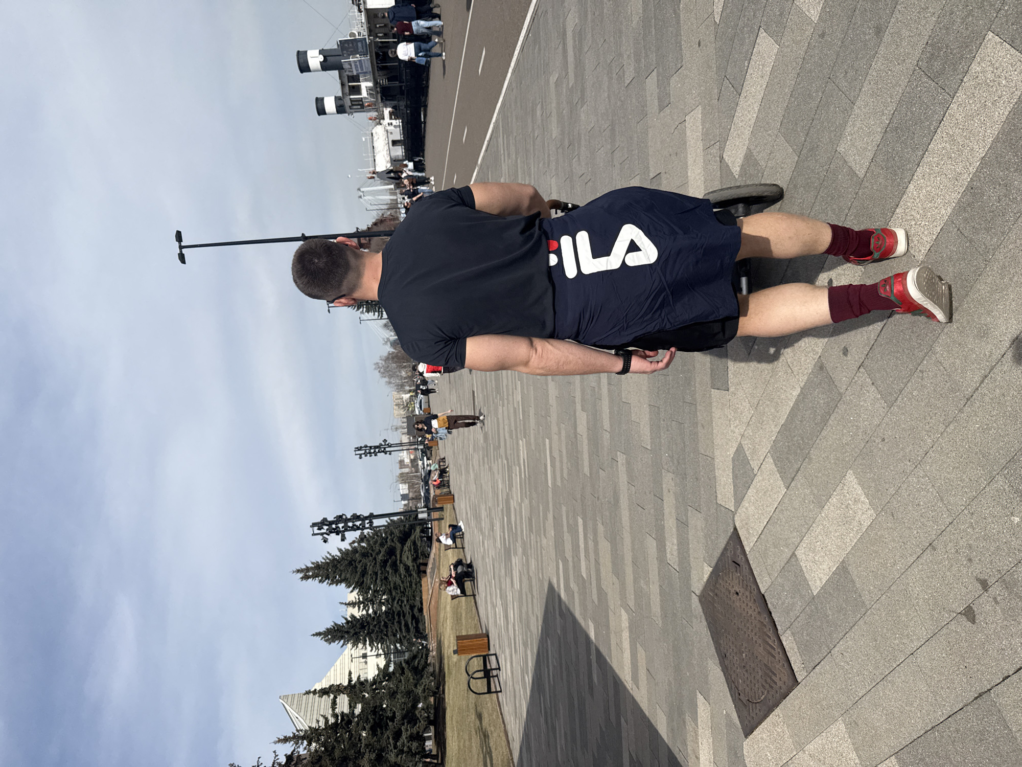A medium-shot from behind of a man with short hair wearing a navy blue t-shirt and shorts with white FILA lettering, burgundy socks, and red shoes is riding a small wheeled device. The man is on a paved area with a patterned design. Behind him, there are trees and light poles. In the distance are other people and a ship docked on the river. The location is likely the embankment of the Yenisey river in Krasnoyarsk, Russia, based on the architecture and ship type.