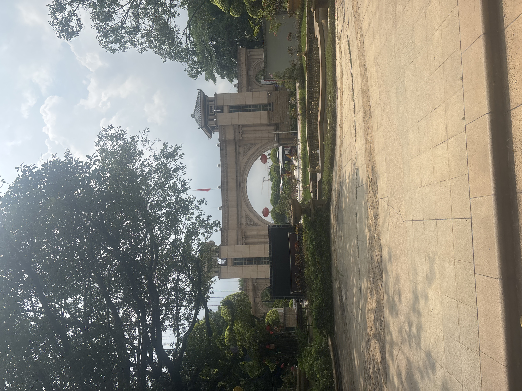 The image captures an arched gateway in a park-like setting in Nanhai District, China. The gateway is constructed of light-colored stone, with ornate detailing and a prominent tower-like structure on the right side. A red flag flies from a flagpole atop the gateway. In front of the gateway, there is a landscaped area with trees, bushes, and a dry fountain. A black rectangular sign is positioned in the foreground, partially obscured by foliage. The ground in the foreground is paved with large, square tiles, with shadows from overhead trees creating a dappled effect. The sky above is partly cloudy, with bright sunlight illuminating the scene. Red lanterns are visible hanging from trees on the left side of the image and near the gateway. In the background, there are glimpses of what appears to be a road with some vehicles and more trees. The overall impression is of a well-maintained public space with traditional architectural elements.