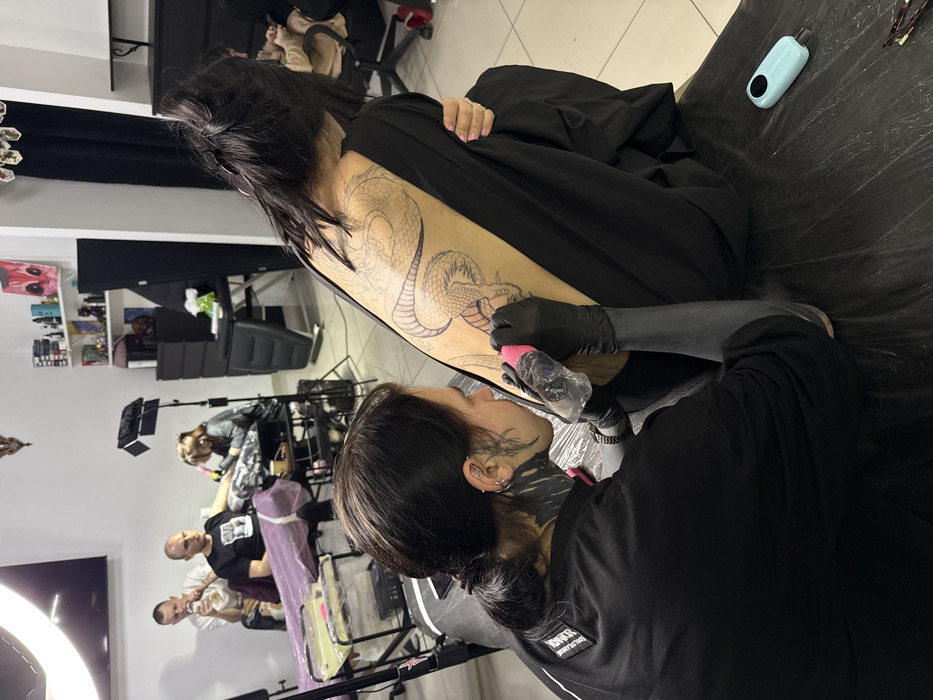 This image depicts the interior of a tattoo studio in Rostov-on-Don, Russia, with multiple tattoo artists at work. In the foreground, a tattoo artist with dark hair tied back, extensive black-out tattoos on the neck, and multiple ear piercings, wearing a black t-shirt and black gloves, meticulously tattoos the upper back of a female client. The artist holds a pink-sleeved tattoo machine, working on the outline of a large serpent or dragon design. The client is seated, facing away from the viewer, with her dark hair loosely clipped up, exposing her back. She wears a loose black garment. The table in front of her is covered with a dark plastic sheet, featuring a light blue, portable fan or similar device to her right.
In the mid-ground to the left, a second tattoo artist, wearing a light-colored shirt, is visible tattooing the forearm of a male client with a shaved head, who is seated on an examination chair draped in purple. A large ring light stands prominently next to this setup. Further in the mid-ground, a third tattoo artist with red hair is seen operating a tattoo machine on another client, whose details are partially obscured. A fourth individual is seated in a dark chair or booth to the right.
The studio features white walls, a light-colored floor, and dark curtains along one wall. Shelves on the rear wall display various art pieces, figurines, and bottles. Professional tattoo equipment, including additional portable lights, tables, and storage units, is visible throughout the brightly lit space.