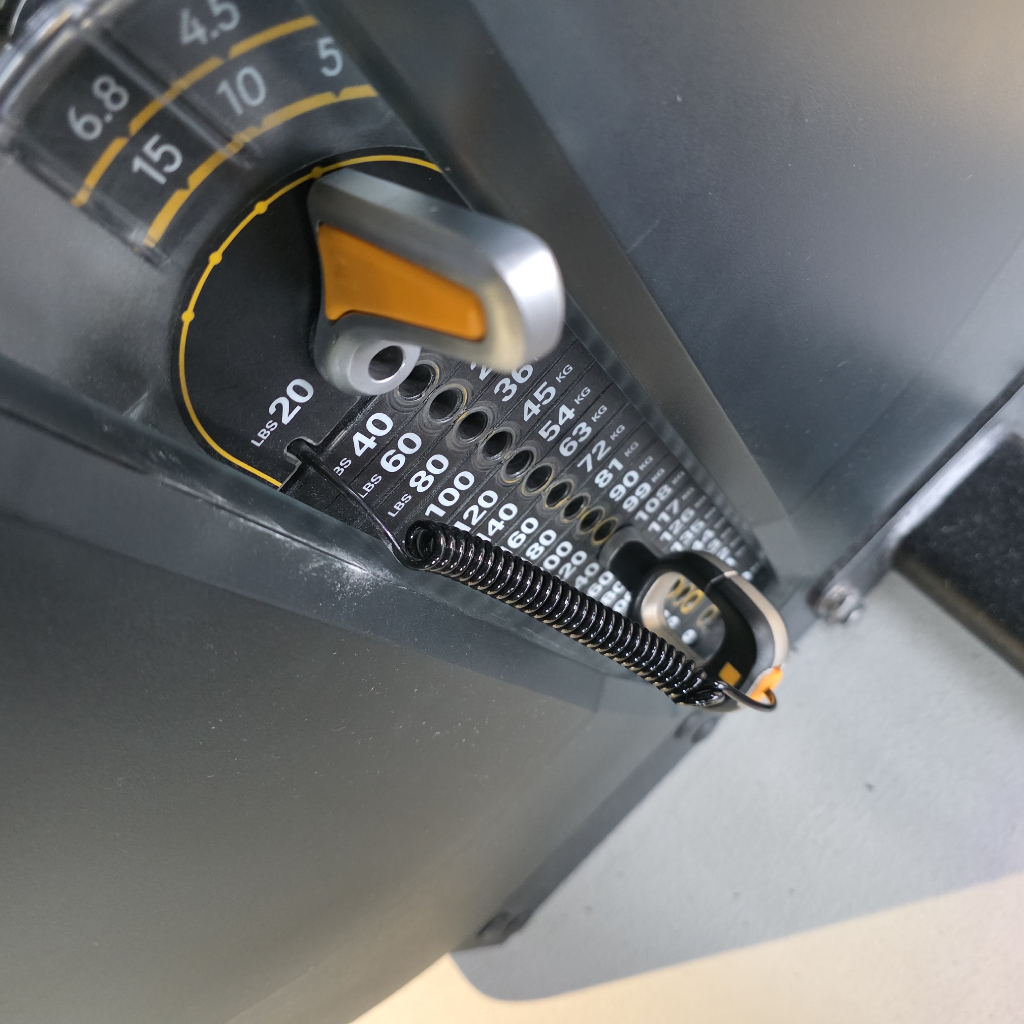 This close-up image depicts the weight selection mechanism of a fitness machine, likely in a gym setting. The central focus is a grey and orange handled pin inserted into a weight stack, which is attached by a coiled black spring. The machine's surface is predominantly dark grey and black, with bright yellow accents highlighting measurement scales.
No people or direct activity are visible, but the setup suggests an exercise machine ready for use, with a weight increment selected. The lighting appears to be artificial, typical of an indoor environment. The surfaces show some signs of use, including minor dust or scuff marks.
Various weight measurements are visible in both pounds (LBS) and kilograms (KG):
- **Pounds:** "LBS 20", "LBS 40", "LBS 60", "LBS 80", "100", "120", "140", "160".
- **Kilograms:** "36 KG", "45 KG", "54 KG", "63 KG", "72 KG", "81 KG", "90 KG", "99 KG", "108", "117", "126", "135".
Additionally, there's a rotary dial or indicator with numbers "6.8", "4.5", "15", "10", "5", possibly representing fine adjustments or another unit of measurement.