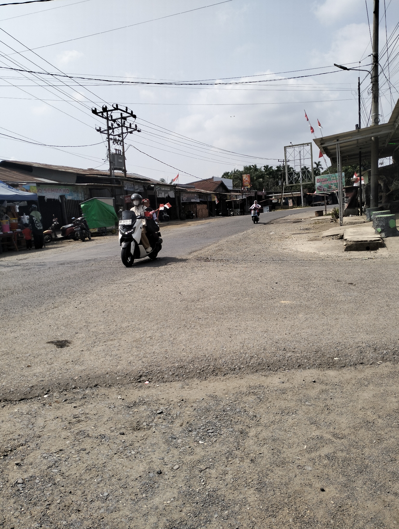 The image depicts a street scene in Serdang Jaya, Indonesia, on a bright, cloudy day. The road is paved with asphalt and appears to be in fair condition, with some gravel and debris present. On the left side of the road, there are several small buildings or shops with corrugated metal roofs, and awnings. A blue tent is set up in front of one of the shops, and several people, including a child, are visible near it. Various motorcycles are parked along the roadside.

In the middle of the road, a person wearing a helmet and a mask is riding a white scooter. The scooter has a license plate that reads "BR 3413 EB". Another person on a scooter is visible further down the road. On the right side of the road, there is a covered walkway or structure, possibly part of a business. Several Indonesian flags are visible on flagpoles. Overhead, a complex network of power lines and poles crisscrosses the sky. The background shows a line of trees and more buildings. The overall impression is of a typical street in a developing urban or semi-urban area.