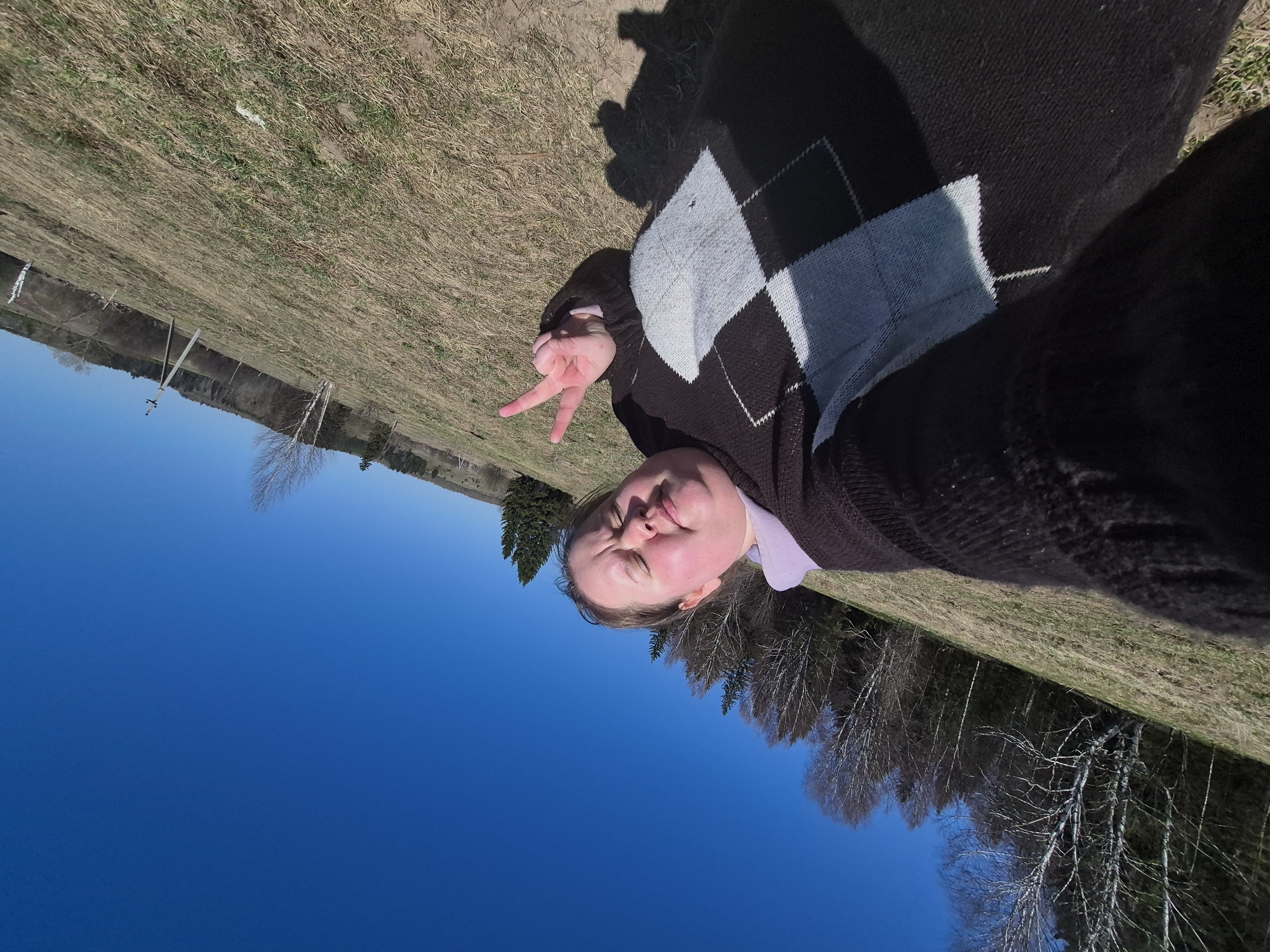 The image shows a woman lying on the ground outdoors in Дойная, Russia. She is wearing a dark brown argyle sweater and a lavender shirt. Her right hand is raised, making a peace sign. The ground is covered in dry grass. In the background, there are trees, a blue sky, and a fence. The image appears to be a selfie taken with a wide-angle lens.