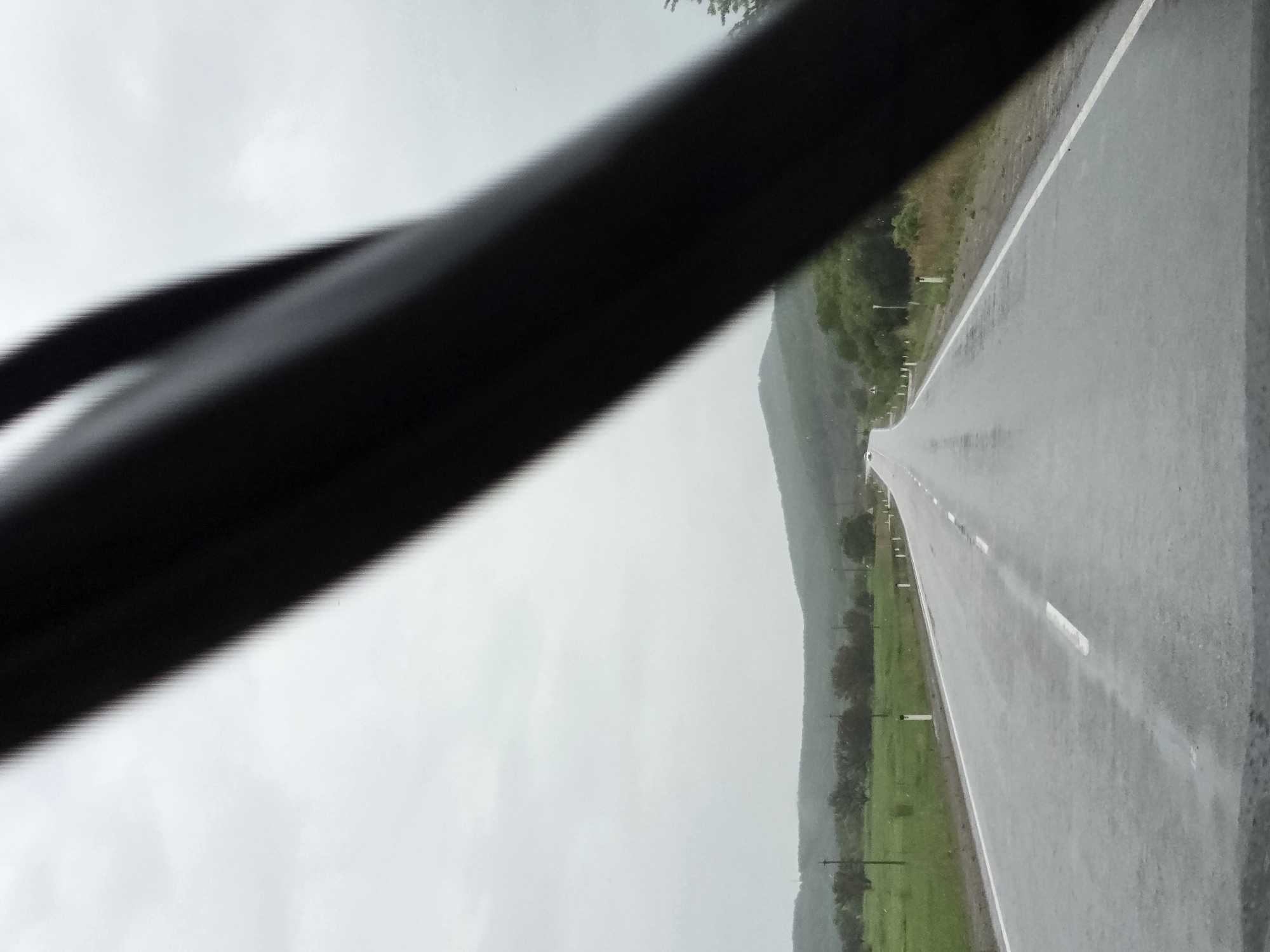 A view from inside a vehicle captures a long, wet asphalt road stretching into the distance under an overcast sky. A dark, blurred object, likely a car dashboard or a pillar, diagonally crosses the foreground from the upper left to the lower right, partially framing the scene. The road, marked with white dashed lines, appears recently rained upon, reflecting the muted light of the sky. Green fields and low-lying vegetation line both sides of the road, with some small posts or markers visible alongside. In the mid-ground, a range of low hills or mountains extends across the horizon, blending into the uniformly gray, cloudy sky. There are no people visible, and the road appears empty. The overall impression is one of a journey through a rural or semi-rural landscape during an overcast and wet day. The location context suggests this scene is in Бабстово, Russia. No discernible text is present in the image.