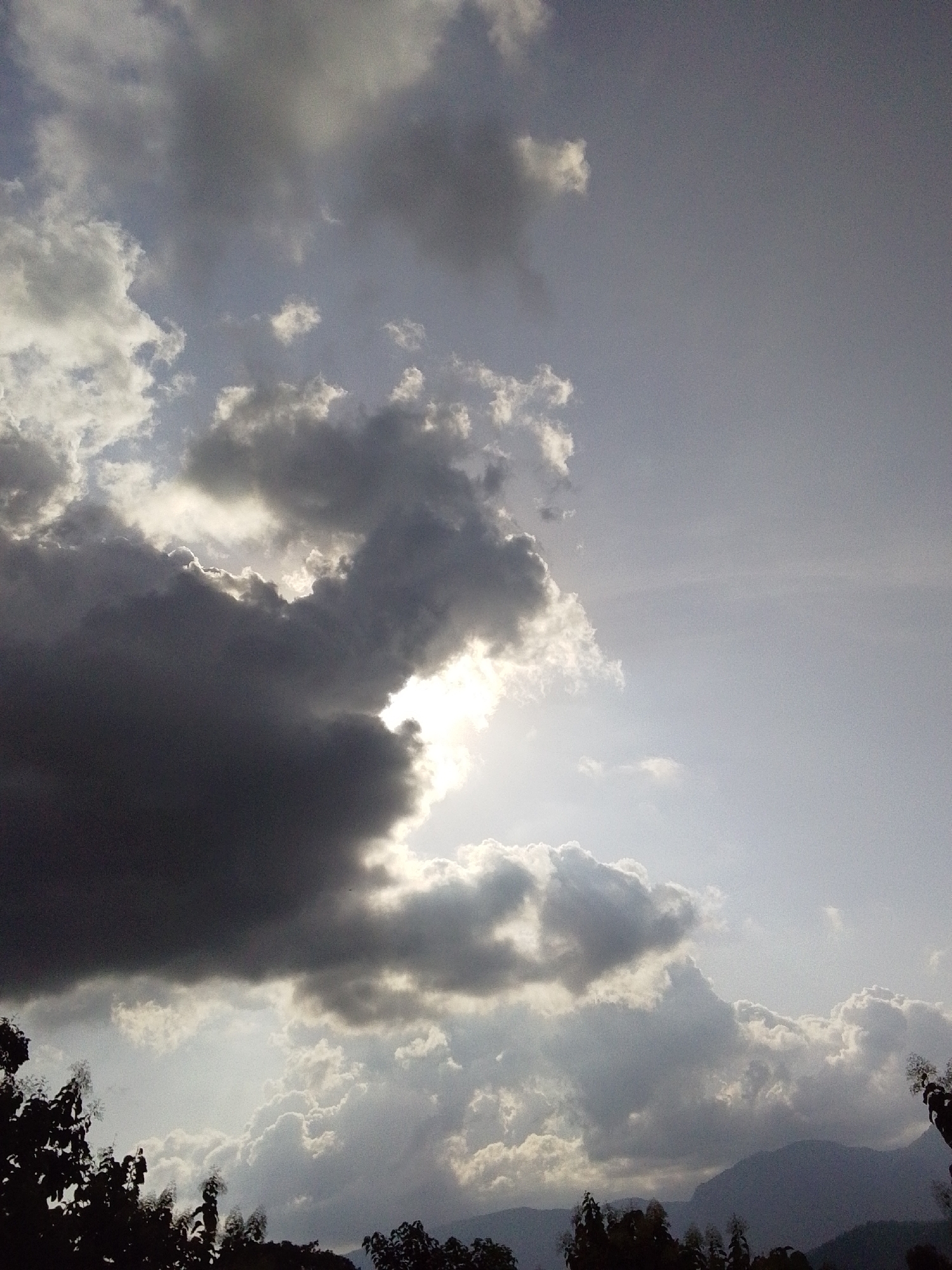 The image shows a dramatic sky with heavy, dark clouds interspersed with areas of bright sunlight. In the foreground, silhouetted trees are visible against the sky, suggesting a natural or semi-natural environment. The clouds are textured and appear to be moving, with some sunlight breaking through in patches, creating a sense of depth and atmosphere. The overall mood is somewhat moody and overcast, with a hint of a storm possibly passing or approaching. There are no people or specific identifiable landmarks visible to pinpoint a location within Serti, Nigeria, but the presence of trees and what appear to be distant hills suggests a landscape outside of a dense urban area.