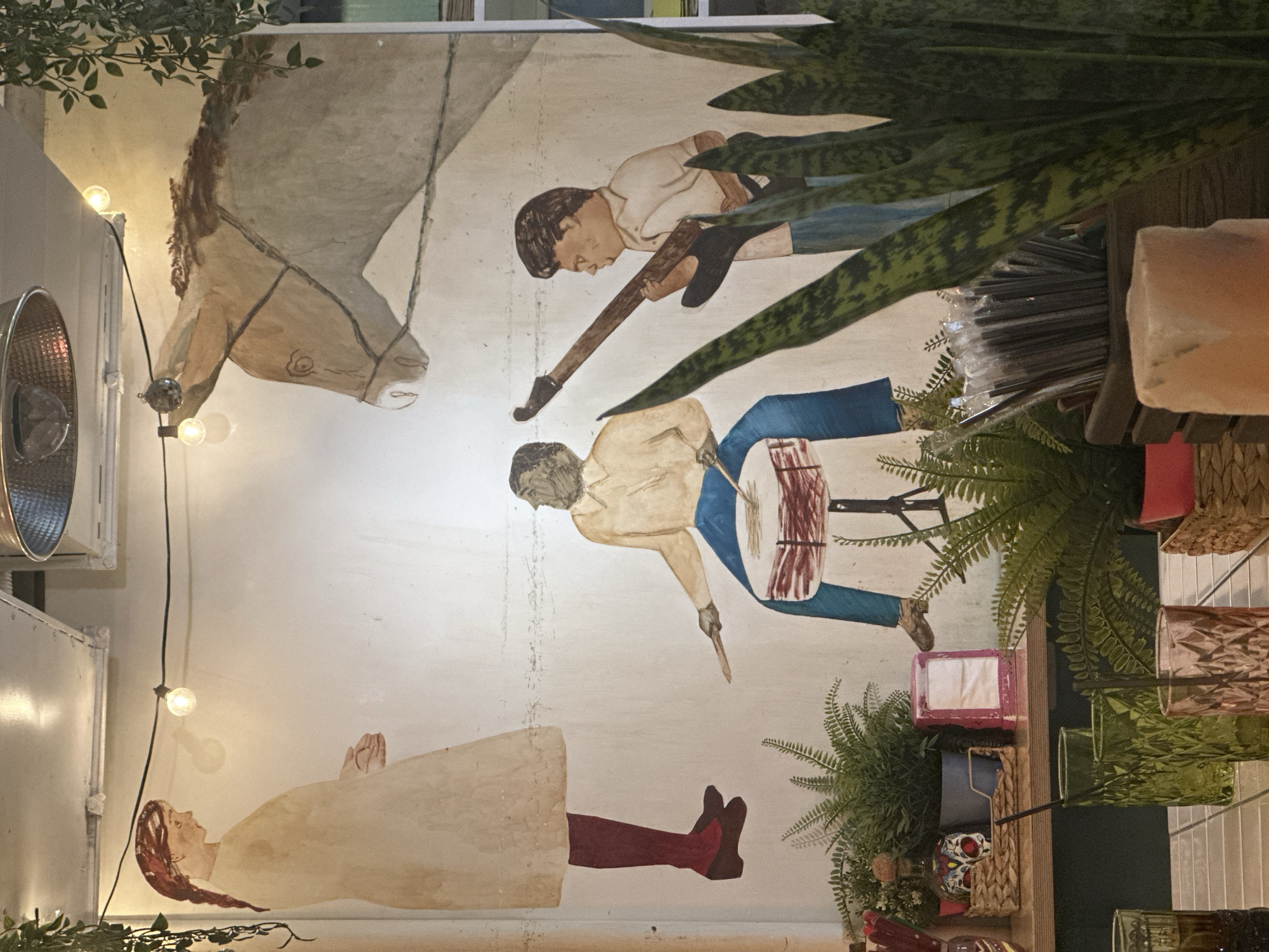 The image captures an interior wall adorned with a large, rustic mural and various decorative elements, likely within a cafe or restaurant setting in Novosibirsk, Russia. The photograph itself is rotated 90 degrees clockwise, presenting the mural on its side.
The mural, painted in a folk-art style with muted, earthy tones, depicts several figures and a horse. On the upper left, a light-colored horse's head with a simple bridle is shown, looking serenely forward. Below it, a male figure with dark hair, wearing a white shirt and blue pants, leans forward, playing a long, dark string instrument. Further down and to the right, another male figure, also with dark hair, a light shirt, and blue pants, is seated and actively playing a red and white drum with two sticks. On the lower left of the mural, a female figure with reddish-brown hair, dressed in a long, light-colored garment and dark red boots, appears to be lying down or floating, looking upwards with a calm expression.
The wall is illuminated by a string of warm-toned incandescent bulbs, suggesting an evening or indoor setting. Several real green plants, including ferns and snake plants, are integrated into the scene, some hanging from above and others positioned on shelves below the mural, creating a lush overlay. These wooden shelves hold various items: plastic-wrapped bundles (possibly straws), an unlit rectangular object, a pink tissue box, wicker baskets, and several glasses in green and other hues. A decorative skull-like object is also visible on one of the shelves. The overall atmosphere conveyed is cozy, artistic, and culturally rich. No discernible text is visible in the image.