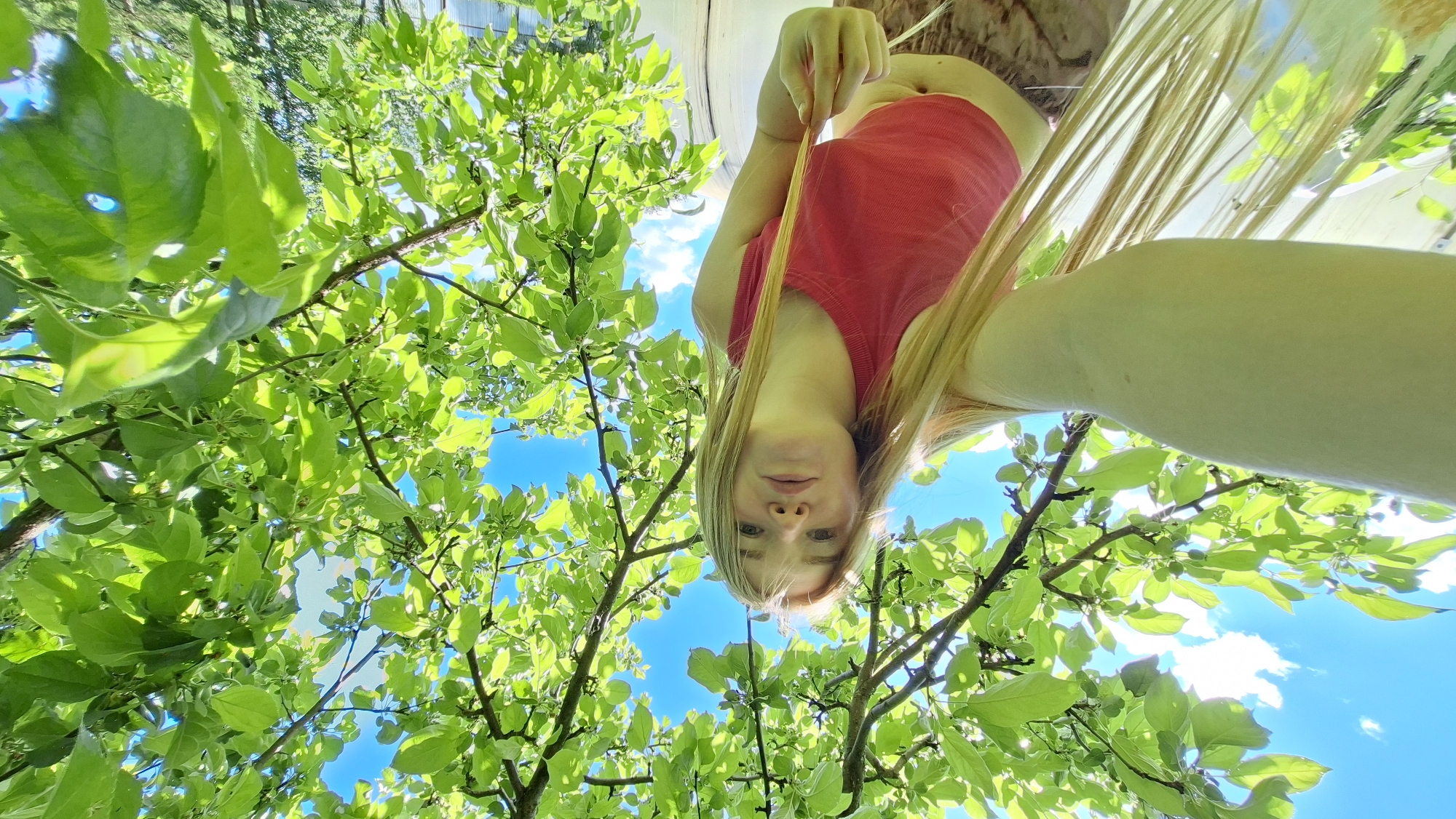 A young Caucasian woman with long blonde hair is captured in a low-angle selfie, taken from beneath a canopy of bright green leaves. She is wearing a pink tank top and patterned bottoms. The background reveals a bright blue sky with scattered white clouds, suggesting an outdoor, possibly daytime setting. The image appears to be taken outdoors, with a focus on the lush foliage of a tree. The location of Большой Букор, Russia is not evident from the image itself.
