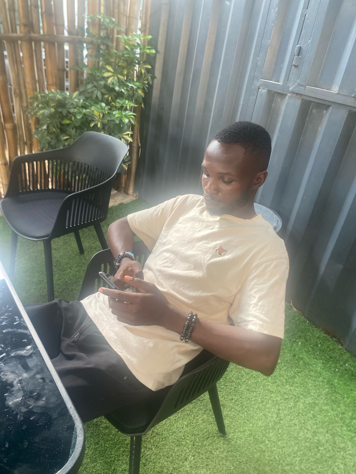 A man sits at a black chair on artificial turf in Barden Yana, Nigeria. He is wearing a light yellow t-shirt with a small insignia on the left chest and black shorts. He has dark skin, a short haircut, and is looking down at his phone, which he holds in his right hand. His left hand rests on his thigh. He wears a beaded bracelet on his left wrist. Behind him is a bamboo wall covered with green foliage. To his right is another black chair and a corrugated metal wall. In the foreground, a black table with a glossy surface is visible.