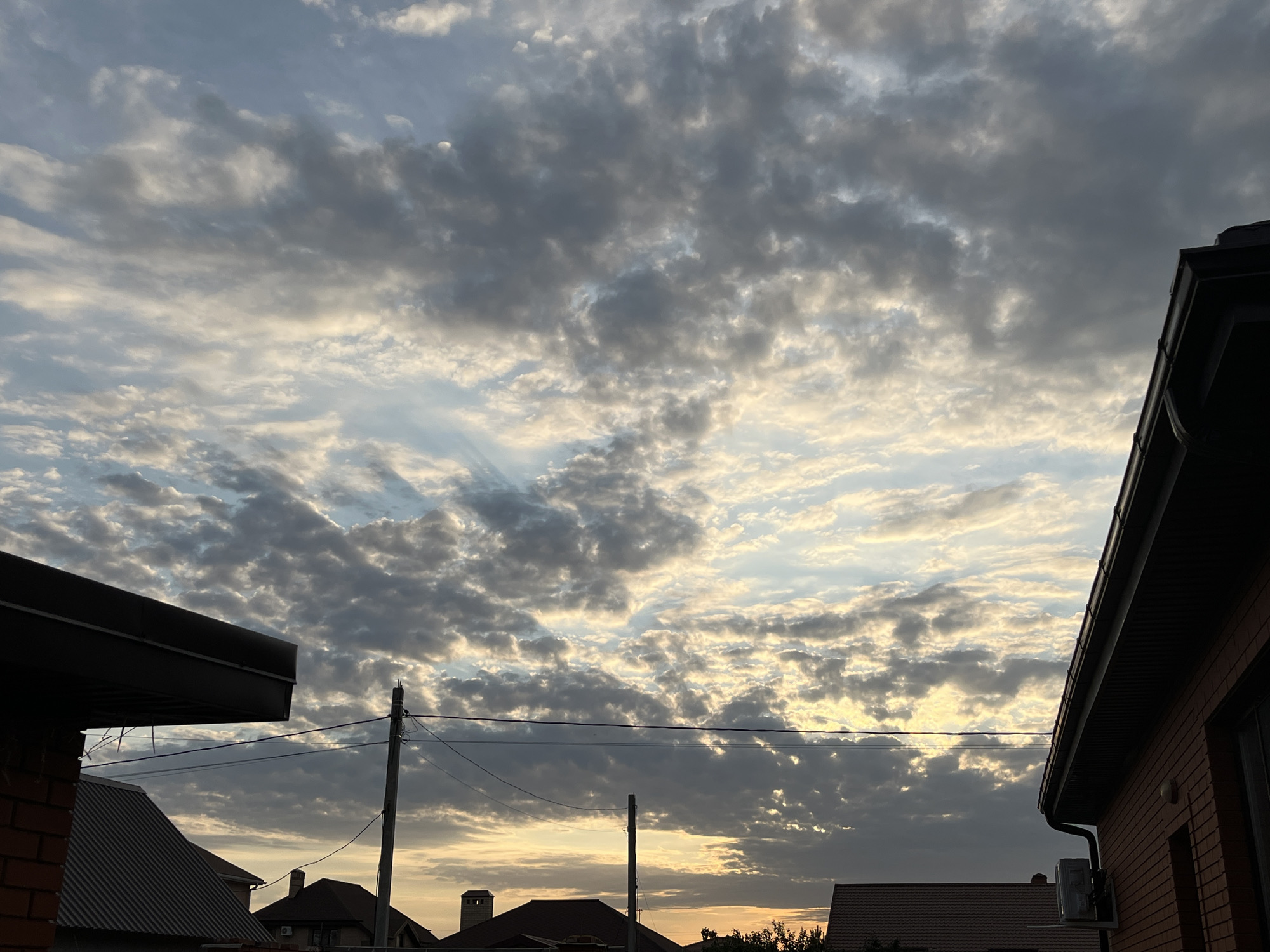 The image shows a cloudy sky over a suburban area, likely in Taganrog City District, Russia. The sky is filled with a mix of dark gray and white clouds, with some patches of blue sky visible. The sun appears to be setting or rising, as evidenced by the warm, yellow light filtering through the clouds. In the foreground, there are several rooftops, a brick building, and utility poles with wires. The overall atmosphere is serene and a bit dramatic due to the cloud formations. There are no people visible in the image.