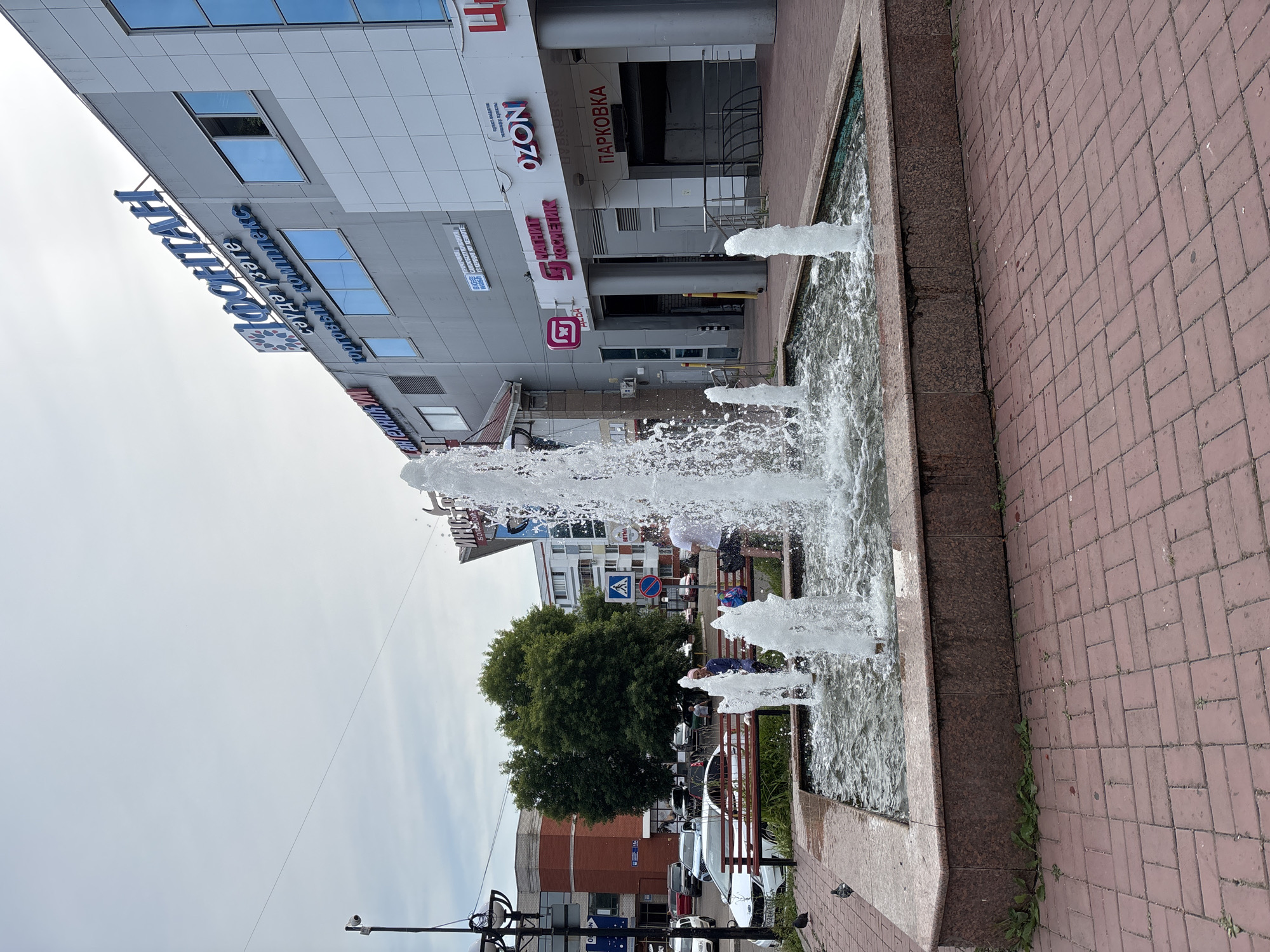 The image shows a public square in front of the "Fontan" shopping center located at 16 G. Tukaya Avenue, Naberezhnye Chelny, Russia. In the foreground, a stone-edged fountain with multiple jets of water is active. The ground is paved with red brick. A few pigeons are visible near the fountain. In the middle ground, a wooden bench is occupied by at least two individuals. One is seated and appears to be wearing a blue headscarf and a dark jacket. Another person is standing behind the bench, facing away from the camera, wearing a white t-shirt and dark shorts. Behind them, trees and other buildings are visible. The "Fontan" shopping center dominates the background with its modern facade featuring large windows and prominent signage in Russian. Other signs on the building indicate businesses such as "SanTEKhNlKUM," "OZON," and "PARKOVKA" (parking). The sky is overcast.