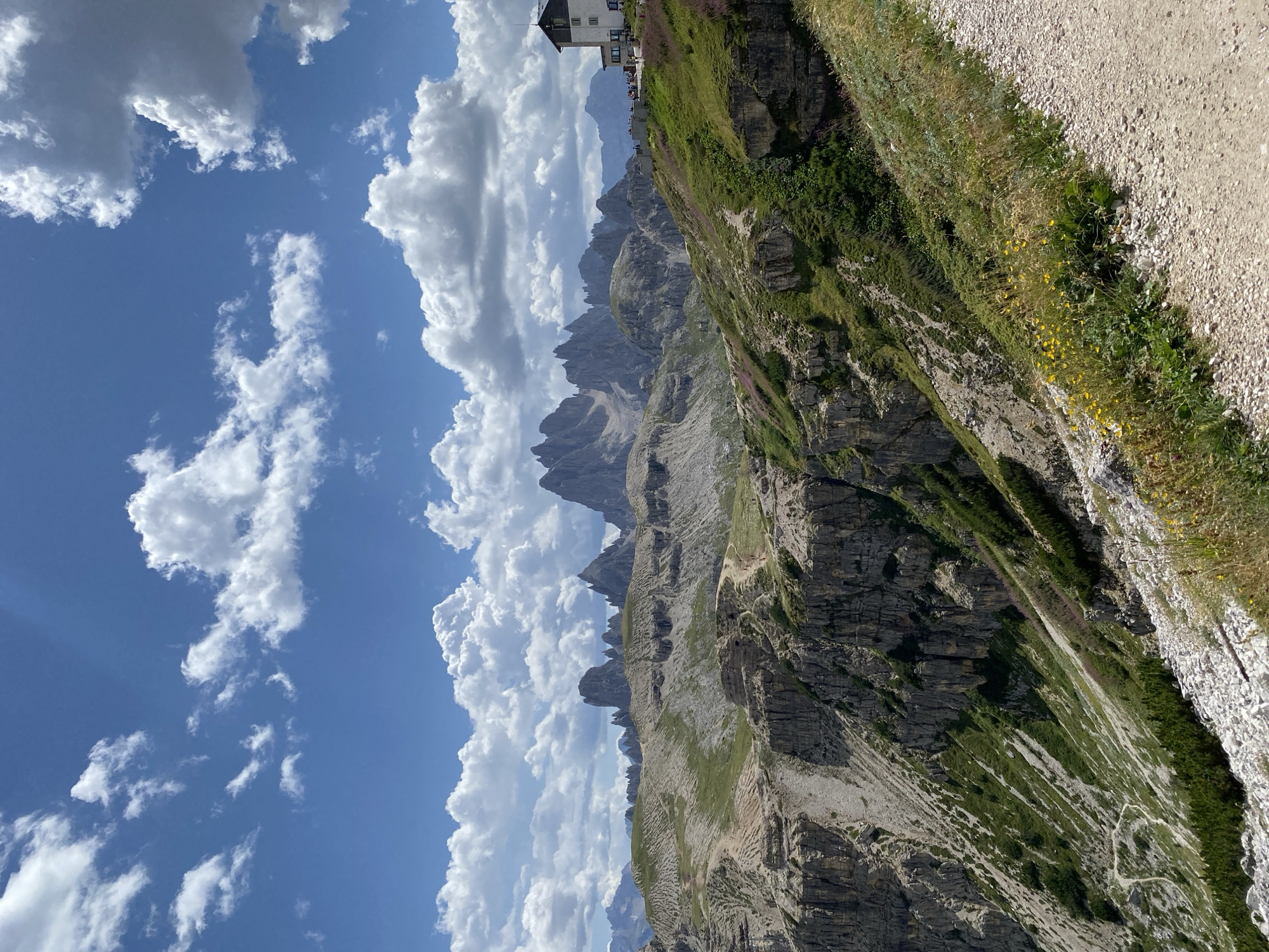 The image depicts a panoramic view of a mountainous landscape, likely the Dolomites, near Auronzo di Cadore, Italy. The foreground features a gravel path winding along a steep, grassy slope dotted with wildflowers and small bushes. Rocky cliffs descend dramatically to the left. In the middle ground, the terrain transitions to rolling, green hills with visible hiking trails. The background is dominated by a rugged mountain range with sharp, jagged peaks under a bright blue sky filled with fluffy white clouds. To the right, a white building with a dark roof, appearing to be a mountain refuge or hotel, sits atop a hill, with a few people visible on its terrace. The overall scene conveys a sense of natural grandeur and outdoor recreation.