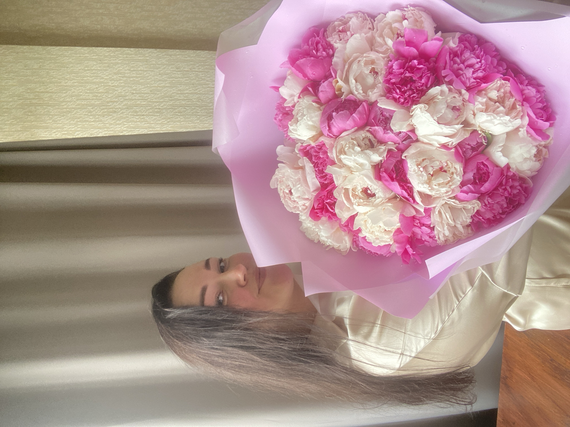 A woman holds a bouquet of pink and white peonies wrapped in light pink paper. She is wearing a light-colored, satin shirt. The background includes a wall with horizontal panels and a draped curtain. The scene appears to be indoors. The city of Sochi, Russia is not discernible from the image.