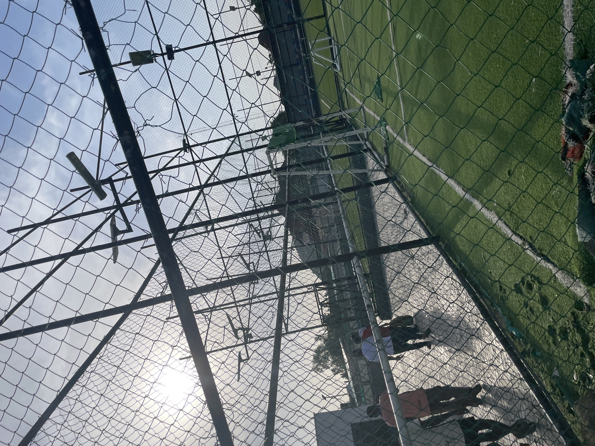 The image depicts an outdoor scene with a focus on a chain-link fence and an artificial turf field. The fence appears to be weathered, with some sections showing signs of damage or wear. Through the fence, a vibrant green artificial turf field with white markings is visible, suggesting a sports facility. In the background, the sky is bright, with the sun prominently shining, indicating it is daytime, likely late afternoon or early morning given the angle of the light.
Several people are visible in the lower right portion of the image, behind the fence. They are dressed in casual attire, including t-shirts and shorts, and appear to be engaged in some activity. Some are standing, while others are seated or interacting with each other. Their presence suggests a community gathering, sporting event, or recreational activity taking place at the facility.
The overall setting seems to be a sports ground or park, possibly in a tropical or warm climate given the clothing of the individuals. The presence of the artificial turf field and the fencing suggests a dedicated area for sports or play. The image is taken from a low angle, looking upwards towards the sky and the fence, which creates a sense of perspective and depth. The light filtering through the fence casts shadows, adding to the visual texture of the scene. No visible text is present in the image.
