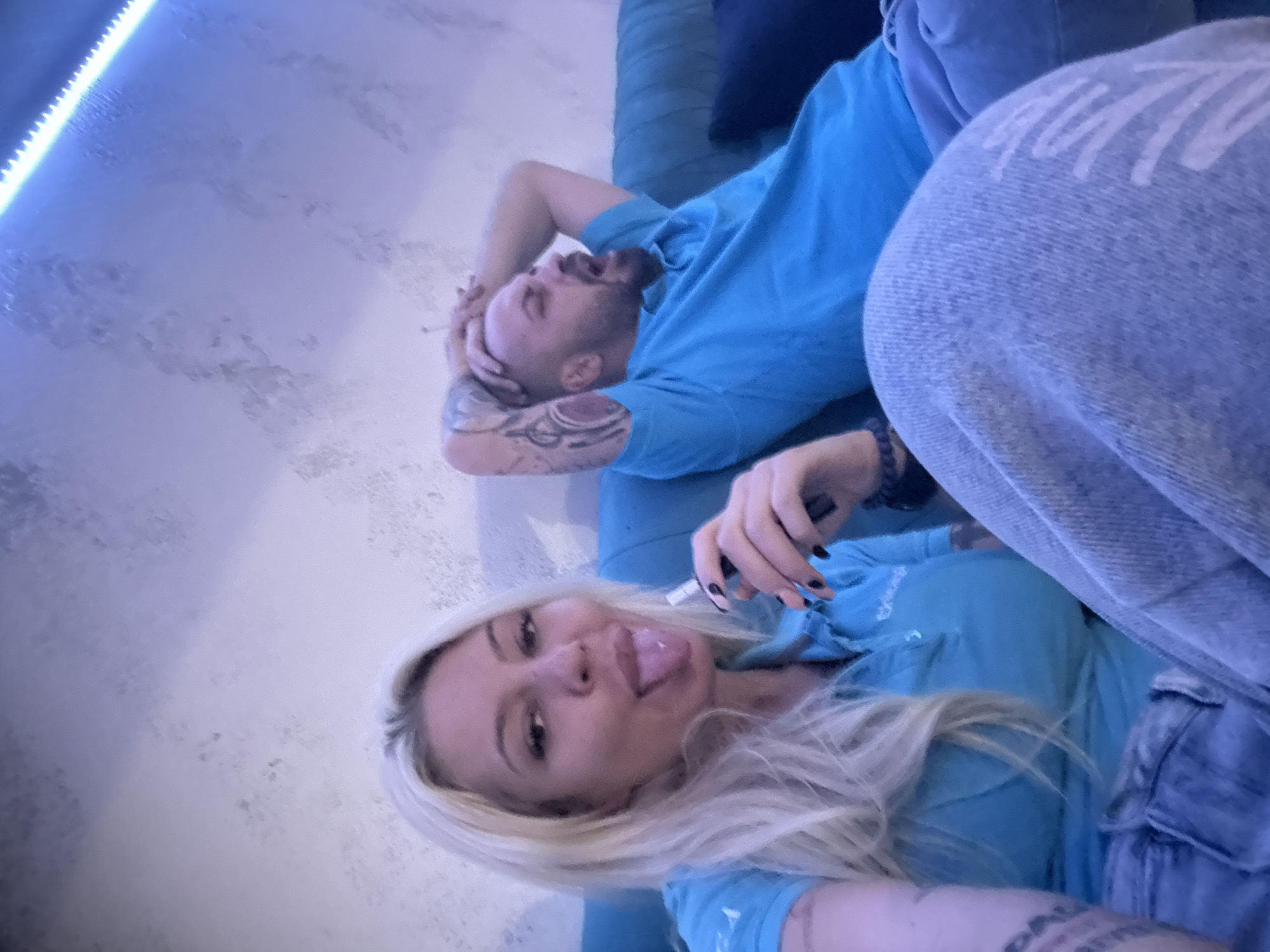 The image, potentially taken in Plovdiv, Bulgaria, captures two individuals, a man and a woman, relaxing indoors on a blue couch or bed. The photograph is composed from a slightly high angle and is tilted counter-clockwise.
The man, positioned in the upper portion of the frame, has a short beard and visible arm tattoos. He wears a blue t-shirt and lies with his hands behind his head, his mouth wide open in what appears to be a yawn or an exclamation. A dark blue pillow is beneath his head.
In the foreground, the woman looks directly at the camera, playfully sticking out her tongue. She has long blonde hair, black nail polish, and wears a matching blue t-shirt and light blue jeans. Her left hand holds a slender, white cylindrical object, resembling a vape pen or cigarette. Tattoos are also visible on her arm. Faint, stylized white lettering, possibly a brand logo, can be seen on her jeans, though it is not clearly legible.
The background features a light gray, textured wall. A prominent blue LED strip light illuminates the left side of the scene, bathing the room in a cool blue-purple glow, suggesting evening or night. The overall mood is casual and informal.