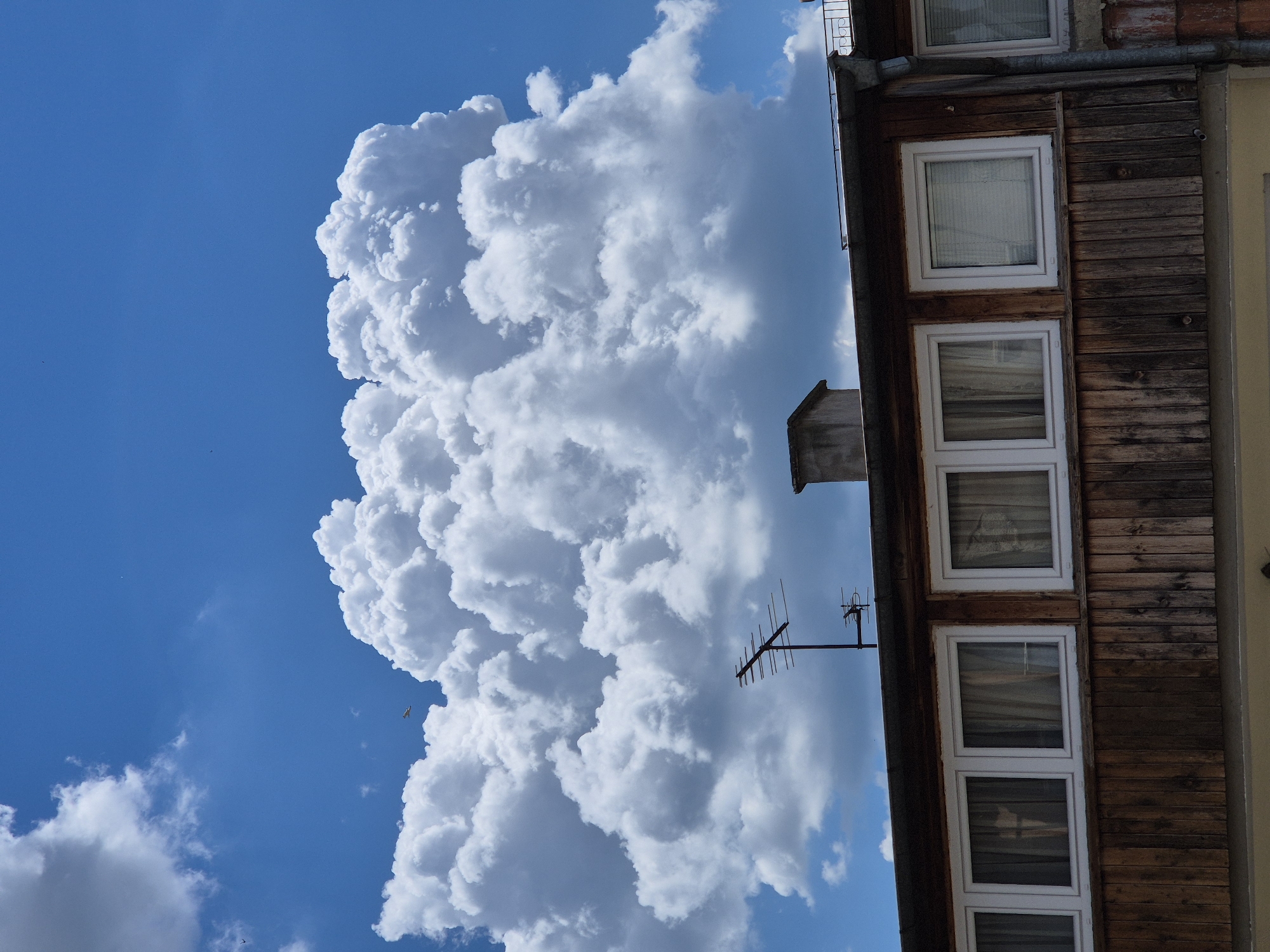 The image shows a view of the sky and the upper portion of a building in Varna, Bulgaria. A large, fluffy white cumulus cloud dominates the upper half of the frame, set against a bright blue sky. Below the cloud, the roofline of a building is visible. The building has a wooden facade with a row of white-framed windows. An old-fashioned television antenna is mounted on the roof, along with a concrete chimney. The building appears to be residential or commercial, with curtains visible through the windows. The overall impression is a sunny day with dramatic cloud formations.