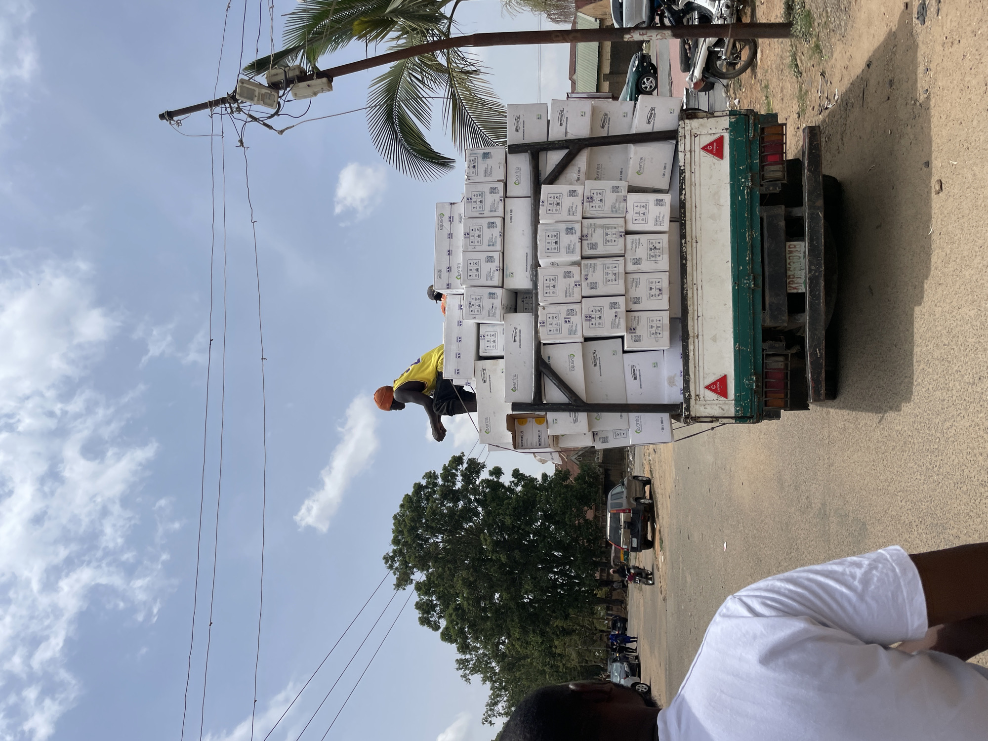 The image, taken in the city of Jos, Nigeria, shows a heavily laden truck on a street. The truck is filled with stacks of white boxes, possibly containing goods for distribution. A person wearing a yellow shirt, dark pants, and an orange hat is on top of the stack of boxes. The truck is parked on a paved road next to a sidewalk. Utility poles with electrical wires are visible in the background, along with trees, other parked vehicles, and people. The sky is partly cloudy. A person wearing a white t-shirt is partially visible in the foreground. The license plate of the truck reads "KRP-630XD".