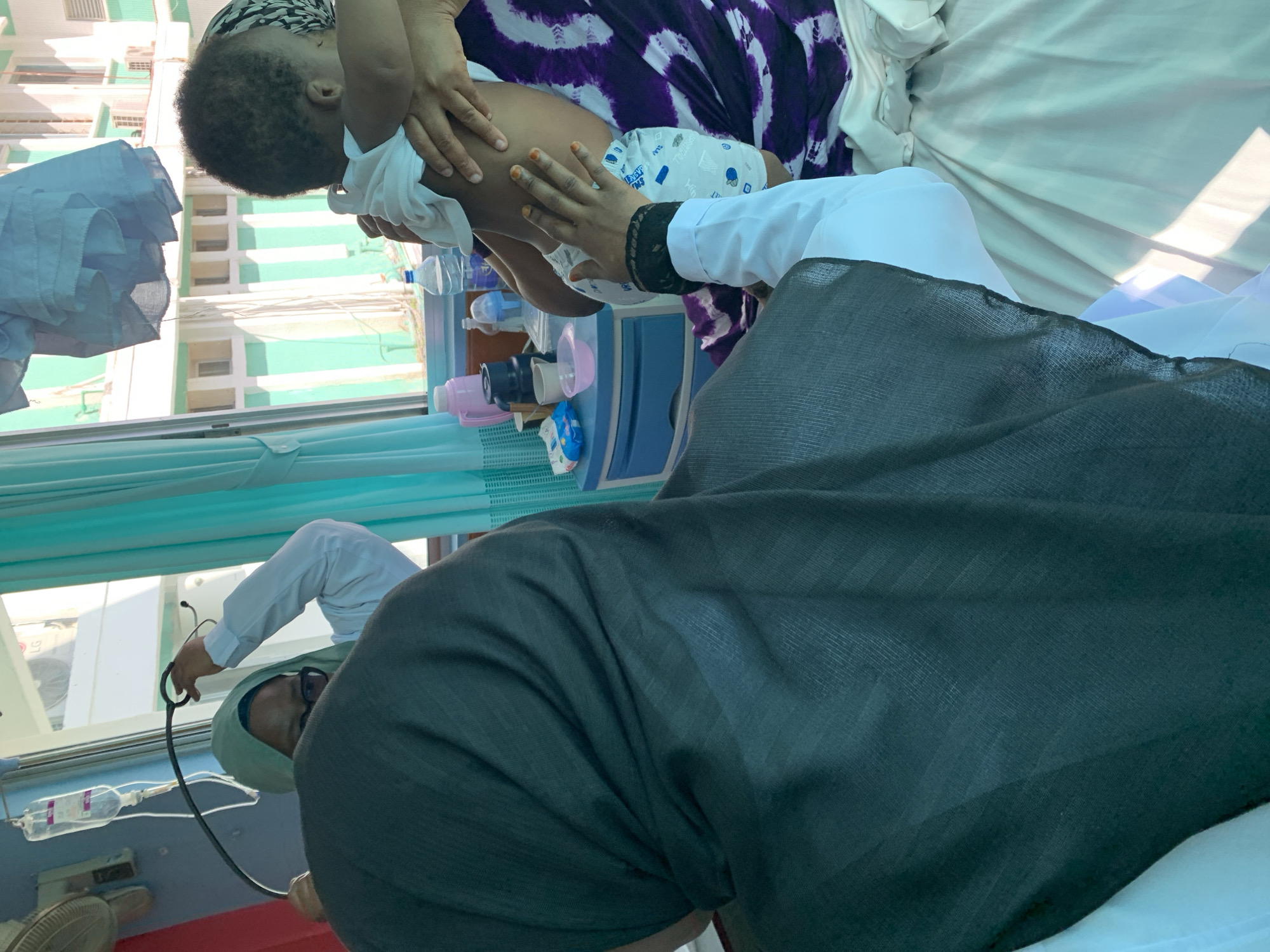 A medical professional wearing a hijab uses a stethoscope on a baby held by someone wearing a purple and white patterned garment. The baby is in diapers. The scene appears to be inside a medical facility, as evidenced by medical equipment and an IV drip visible near a window. Outside the window, there are buildings with light green facades. Based on the setting and architecture, the location is likely Dar es-Salaam, Tanzania.