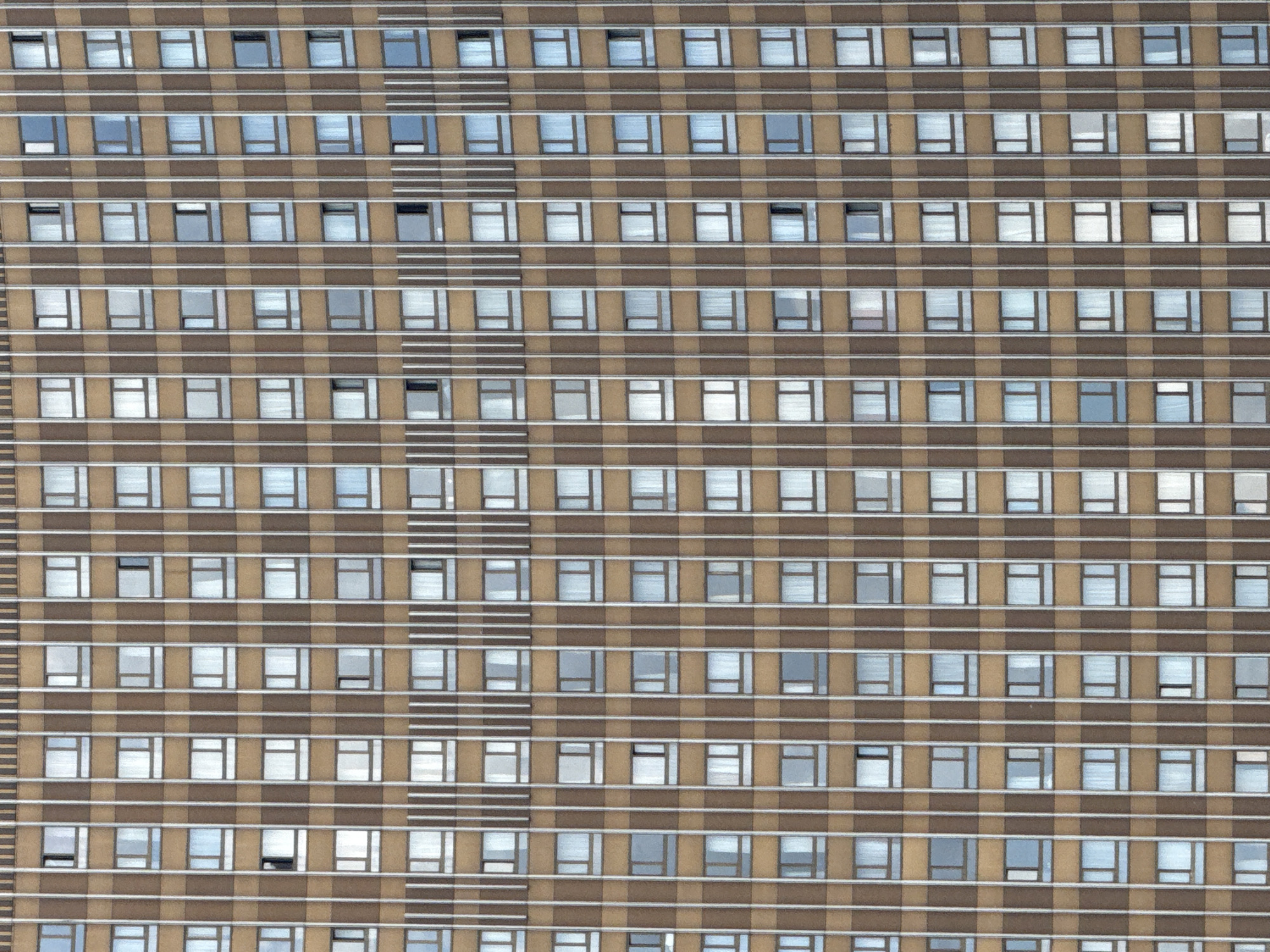 The image shows a high-angle view of a multi-story building with a repeating pattern of windows and horizontal stripes. The building's exterior is primarily brown with light-colored horizontal bands separating each row of windows. The windows appear to be rectangular and have light-colored frames. Some windows reflect light, while others appear darker, possibly indicating different lighting conditions inside. On the left side of the building, there is a vertical element with multiple thin, light-colored stripes. The building is located in Moscow, Russia.