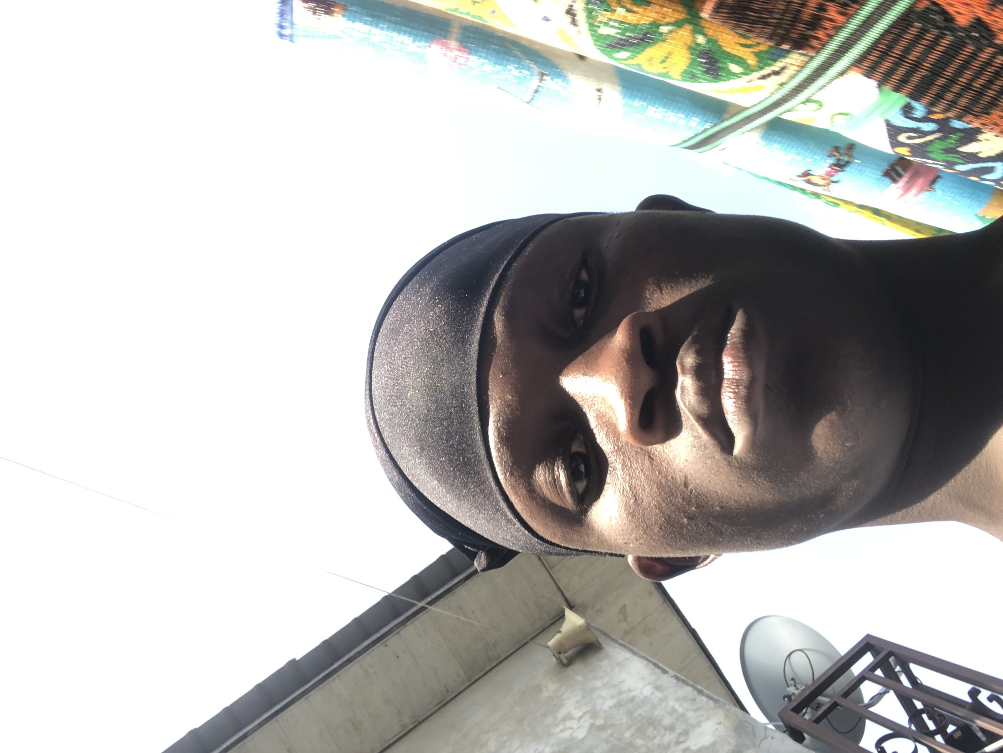An extreme close-up selfie shows the face of a young Black person wearing a black durag, looking directly at the camera. The person's skin is dark and appears to be slightly sweaty, with faint acne marks on the cheeks. Their eyes are dark and open, with their eyebrows faintly visible. The lips are full and appear moisturized. Behind the person, to the left, is a concrete building with a sloped roof and a satellite dish attached to it. To the right, several rolled-up mats with colorful, abstract patterns are visible. The background is a bright, washed-out sky, suggesting it is daytime. The lighting indicates the sun is high and possibly behind the camera, casting shadows that emphasize the contours of the person's face. The composition is a low-angle shot, making the subject appear dominant. The image's composition and the presence of elements like the building and satellite dish could suggest an outdoor setting in an urban or semi-urban environment, potentially in Ado, Nigeria, given the context.
