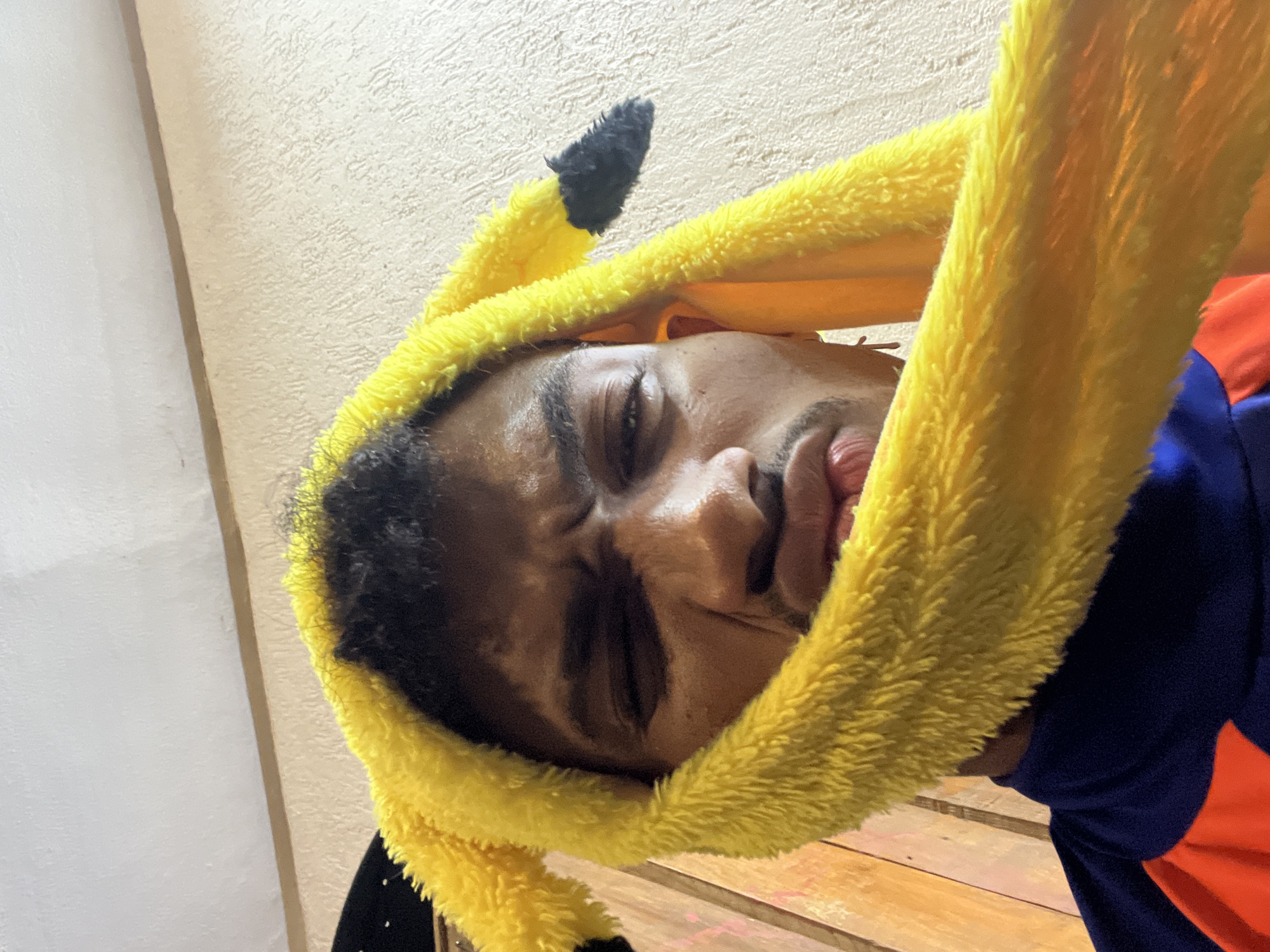A man wearing a Pikachu-themed hood is seen indoors. He has dark hair and appears to be resting or tired, with his eyes partially closed. The background includes a textured wall, and a glimpse of a wooden surface. The image gives no information to identify the city or country where this image was captured.