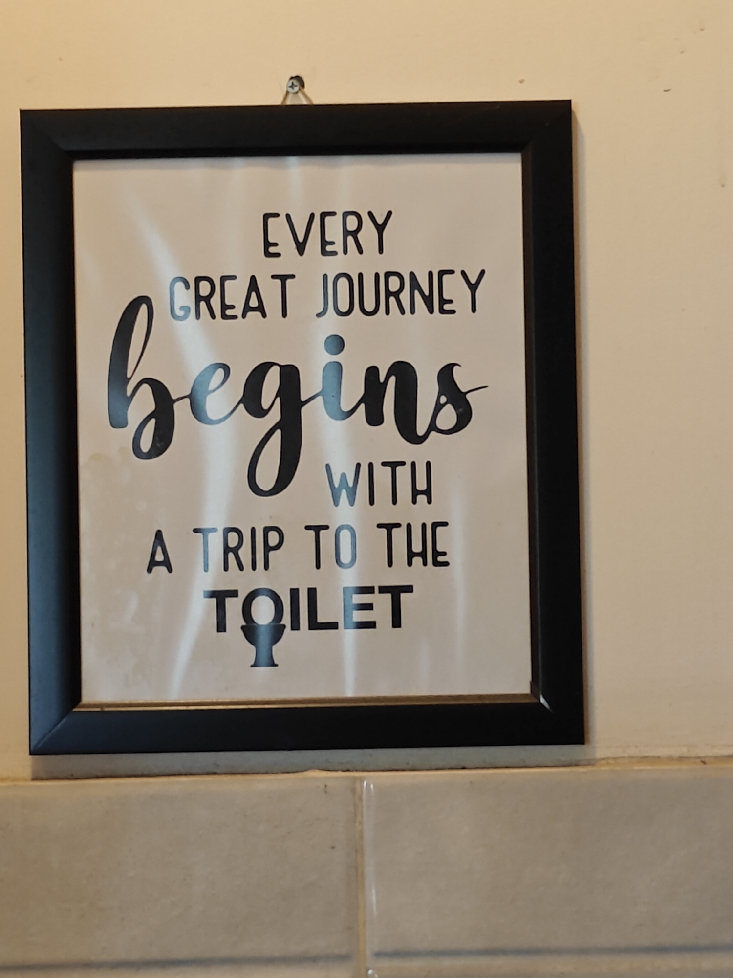 A framed picture with a black border is hanging on a wall. The picture contains text and a graphic of a toilet. The text reads "EVERY GREAT JOURNEY begins WITH A TRIP TO THE TOILET". The picture is hanging from a screw on the wall. The wall appears to be cream-colored. Below the wall with the picture, there is a tiled surface. The scene is indoors, likely in a bathroom. The image is a close-up of the framed picture and its surroundings. There is no discernible information about the city of Ichchhakamana, Nepal in the image.