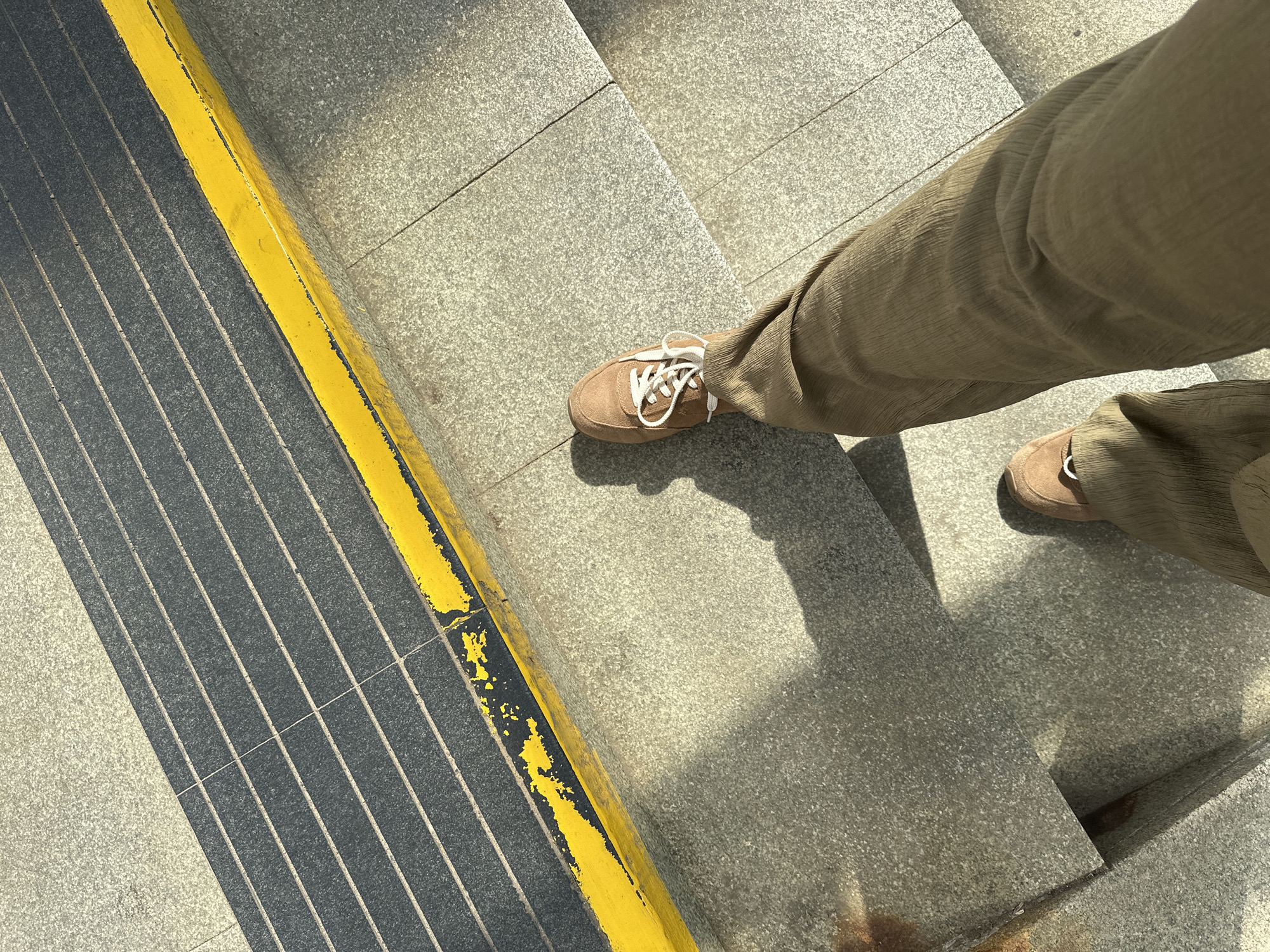 A first-person perspective shot captures a person walking down a set of concrete stairs. The person is wearing brown corduroy pants and brown suede sneakers. The stairs are wide and well-maintained, with a yellow and black safety strip running along the edge of one of the upper steps. The sun casts long shadows across the stairs, indicating it is likely late afternoon or early morning. The surrounding environment appears to be an urban outdoor setting, possibly a public space like a park or a metro station. The image does not contain any specific landmarks or clues that would definitively place it in Minsk, Belarus.