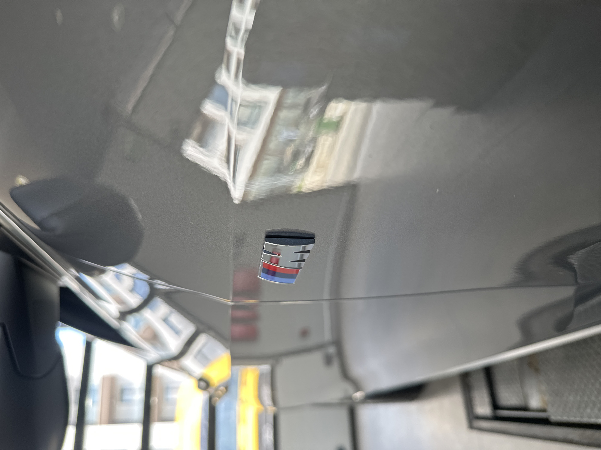 An extreme close-up shot shows the side of a dark gray vehicle. A silver emblem with red, blue, and white stripes is affixed to the car's body. Reflections of buildings and a yellow object are visible on the glossy surface of the car. The car appears to be in an indoor setting, possibly a garage or workshop, as indicated by a metal grate visible in the foreground.