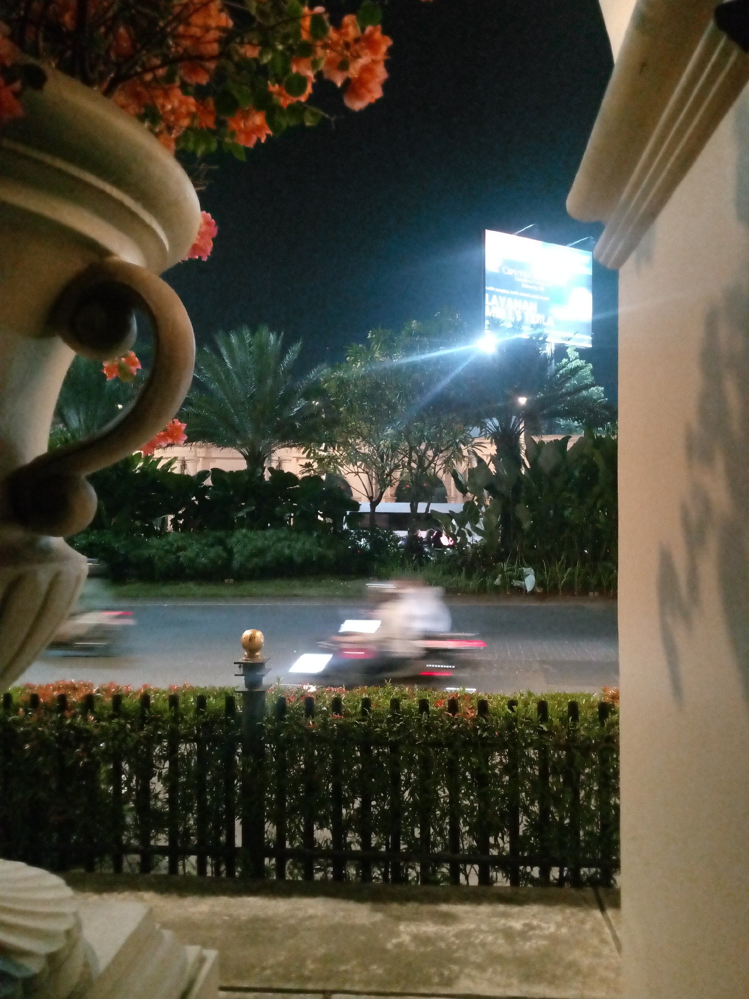 This is a night photograph taken from a ground-level perspective, looking across a street towards a billboard. In the foreground, a large, ornate stone vase with decorative handles is visible on the left, with orange flowers and green foliage above it. A dark green hedge with some red flowers runs horizontally across the lower part of the frame, behind which is a road. The road shows motion blur, indicating vehicles passing by, with streaks of white and red lights suggesting headlights and taillights. A slender black fence with vertical slats and a small golden sphere on top of a post stands in front of the hedge. On the right side of the image, a textured, light-colored wall with shadow patterns is present. In the background, across the street, is a brightly lit billboard displaying text that includes "CARUMBA" and "LAYANAN MILD". Palm trees and other foliage are visible behind the road, and some buildings with arched windows can be seen in the distance, illuminated by the billboard's light. The overall scene depicts a street view at night, possibly in a commercial or residential area, with a focus on the contrast between the illuminated billboard and the dark night sky, and the movement of traffic on the road. The presence of palm trees and the style of the buildings suggest a tropical or subtropical location. Given the context of Bitung Jaya, Indonesia, this scene likely represents a common urban night setting in that region.
