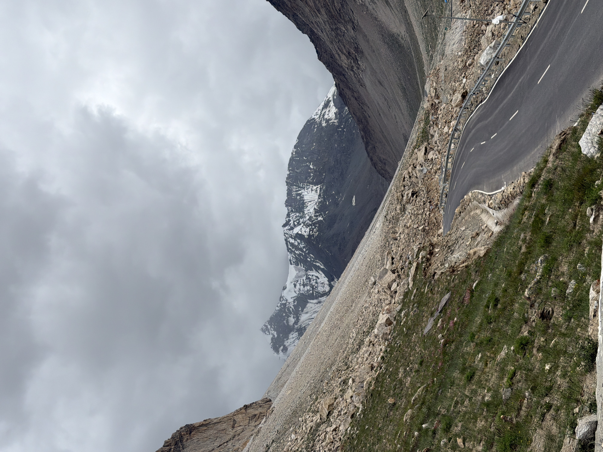 A winding asphalt road curves through a mountainous valley in Darcha, India, under a cloudy sky. The road has white dashed lines marking the lanes and a metal guardrail on the right side. Steep, rocky slopes flank the road, with patches of green grass and scattered boulders. In the distance, snow-capped mountains rise dramatically against the overcast sky. The scene is devoid of people, with the landscape dominating the view.