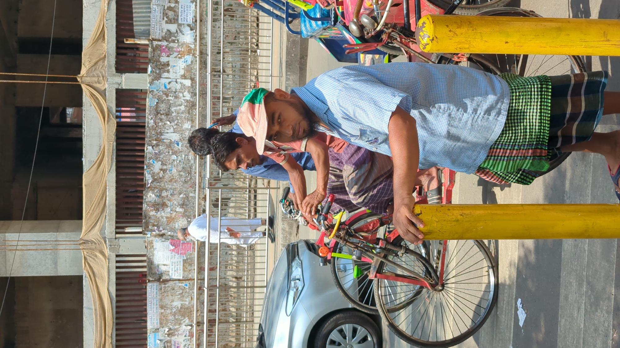 The image shows two men with rickshaws on a street in Dhaka, Bangladesh.
**People:** Two men are visible, both wearing lungis (traditional wrap-around garments). The man closest to the camera wears a blue checkered shirt and a light pink cap. The second man wears a blue shirt and a patterned lungi.
**Subjects/Objects:** The men appear to be rickshaw pullers. Two rickshaws are partially visible. There is a silver car parked in the background. A yellow painted barrier is in the foreground. The background shows a building with what appears to be construction netting and distressed walls.
**Actions/Interactions:** The men are standing next to their rickshaws. The man in the blue checkered shirt has his hands on the yellow barrier. It appears they may be resting or waiting for fares.
**Environment:** The scene is outdoors on a city street. The lighting suggests it is daytime. The general appearance suggests a busy urban environment. The distressed buildings and the presence of rickshaws indicate that this is likely a less affluent area of Dhaka.
