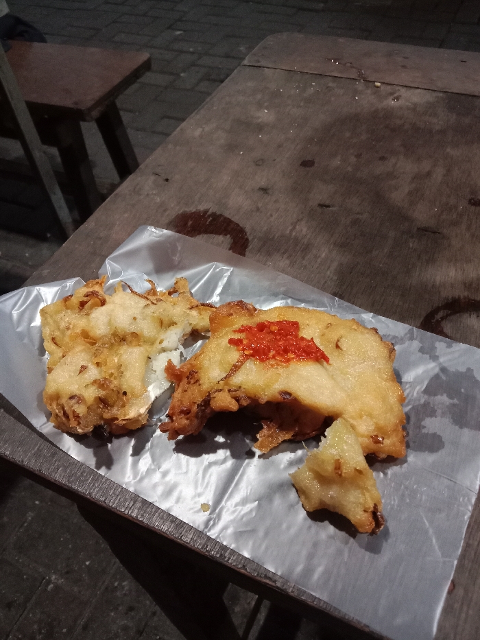 The image shows a plate of fried food on a wooden table. The food is wrapped in clear plastic and is topped with a red chili sauce. There is a wooden bench visible in the background. The coordinates provided correspond to a location in the city of Tugu, Indonesia. It appears to be a street food stall. The scene suggests a casual, informal setting.