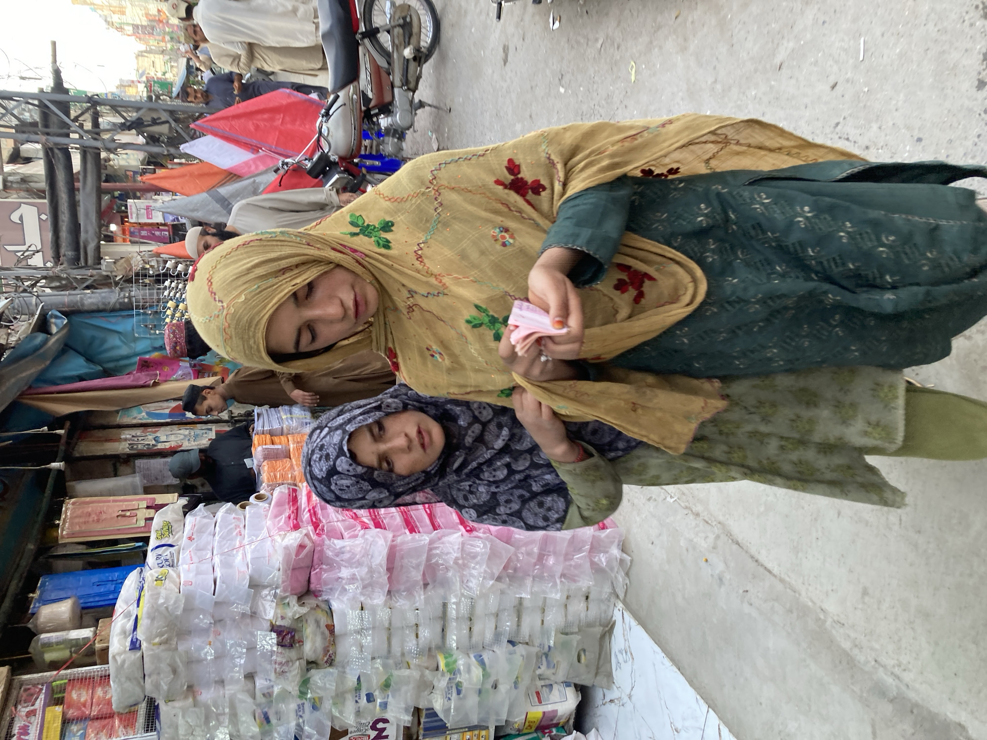 This image captures a vibrant street scene, likely in Quetta City Tehsil, Pakistan, during daylight hours. The central subjects are two girls, an older one and a younger one, positioned in the foreground.
The older girl, on the right, wears a mustard-yellow headscarf (dupatta) with red and green floral embroidery, and a dark green shalwar kameez. She holds several pink Pakistani rupee notes in her right hand. Her gaze is directed slightly downwards or towards the camera, with a gentle, somewhat solemn expression. Her left arm is around the younger girl.
The younger girl, on the left, wears a dark patterned headscarf, possibly in black and white, and light green clothing. Her expression is also subdued, with her eyes slightly downcast. She is positioned slightly behind and to the left of the older girl.
They are standing near a market stall, which is heavily stocked with numerous items, predominantly clear plastic bags containing various goods, possibly small packaged items or other plastic products. In the background, other market stalls are visible, characterized by colorful awnings in red, orange, and white, suggesting a bustling outdoor market environment. Several men, dressed in traditional Pakistani attire including shalwar kameez and prayer caps, are seen among the stalls and walking on the street. Parts of motorcycles are also visible, indicating a busy urban thoroughfare.
The overall scene conveys a snapshot of daily life and commerce in a South Asian street market. No specific, clearly decipherable text is visible on signs or packaging, though some unreadable script can be observed on some items. The emotions conveyed by the girls are quiet and thoughtful amidst the busy market backdrop.