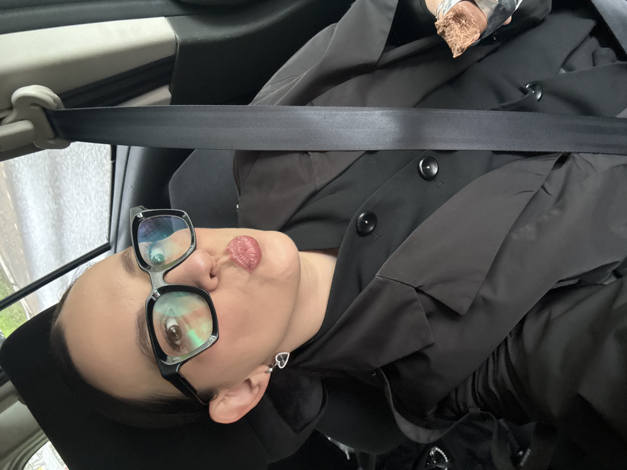 In the interior of a car, a woman with dark hair and glasses is seated in the passenger seat, wearing a black blazer. She is making a kissing face towards the camera and holding a piece of a chocolate bar in her right hand. The seatbelt is fastened across her chest. The car appears to be parked or moving slowly, as indicated by the blurred background which suggests an outdoor environment with some greenery and a road. The exact location within Chaikovsky, Russia, is not discernible from the image.