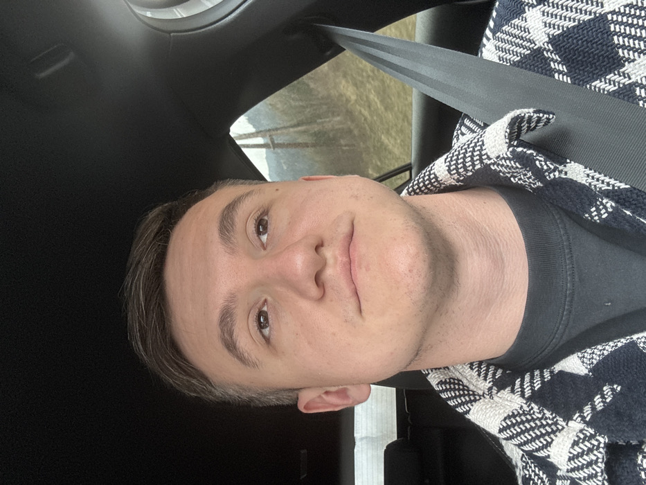 The image depicts a young adult male seated in the front passenger position of a vehicle, located in Murau, Austria. The subject has short, dark brown hair, light skin, and is looking upwards and slightly to the right of the frame with a neutral expression. He is wearing a dark-colored t-shirt beneath a black and white patterned, possibly houndstooth or plaid, long-sleeved shirt. A black seatbelt is diagonally secured across his chest, originating from the upper right of the frame. The vehicle's interior features a dark headliner and trim. Through the vehicle's side window, a mountainous or hilly landscape is visible, characterized by sloped terrain with sparse, brownish-green vegetation and a collection of bare trees. A prominent wooden utility pole stands in the middle distance of the outdoor environment. The scene is illuminated by natural light, consistent with an outdoor setting.