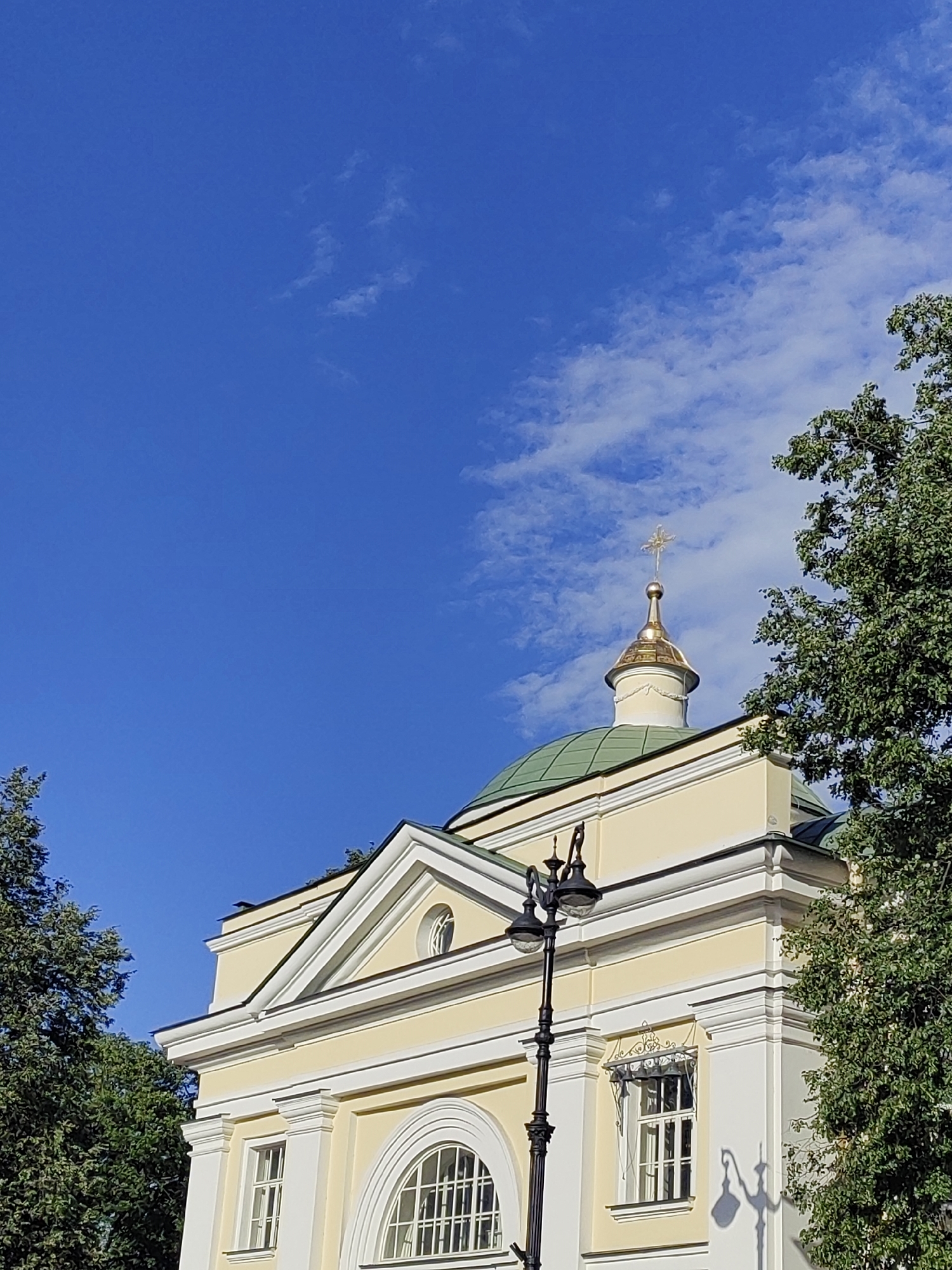 This media file captures the exterior of a yellow building with white trim, featuring a prominent green dome topped with a golden cross. The building appears to be a church or religious institution. To the right and left of the building, lush green trees are visible, their leaves obscuring parts of the structure. The sky above is a vibrant, clear blue, dotted with a few wispy white clouds. A black streetlamp with two globes stands in front of the building. The sunlight suggests it is daytime, likely morning or afternoon, and the clear sky indicates fair weather. The architectural style of the building, with its dome and cross, along with the presence of trees and the characteristic blue sky, are consistent with a location in Saint Petersburg, Russia. No people are visible, and no text is present in the image.