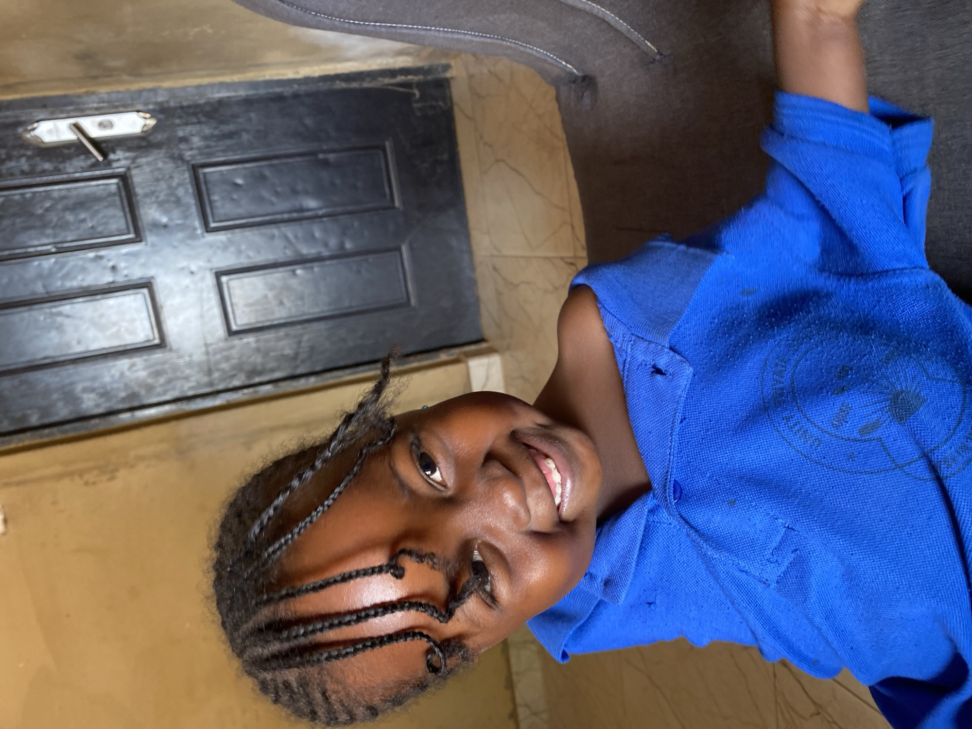 A young Black girl, appearing to be in elementary school, smiles brightly and looks directly at the viewer in this portrait, set indoors in Kuru Dungus, Nigeria. Her head is tilted, and her mouth is open, revealing her teeth, conveying a joyful and friendly expression. She has dark skin and her hair is styled in numerous neat, thin braids or cornrows.
She is wearing a bright blue polo shirt with a collar. The fabric of the shirt shows signs of wear, including some visible pilling and small holes, suggesting it might be a well-used uniform. A faint, dark circular emblem with some text, including the word "UNITY," is visible on the right side of her chest.
The background consists of a dark, paneled door with a simple metal latch mechanism visible on its surface, positioned behind and to the left of the girl. Below the door is a light brown or tan-colored surface, possibly a floor or lower wall. To the upper right, a portion of a gray, upholstered object, possibly a sofa or chair, is visible. The lighting appears to be ambient indoor light. The entire image is rotated approximately 90 degrees clockwise.
