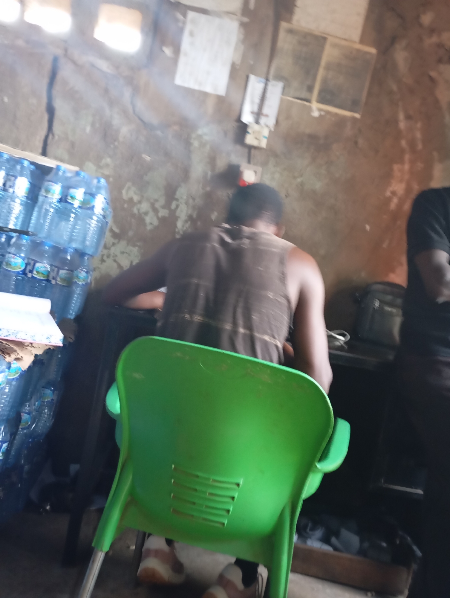 The image depicts an indoor scene, likely a shop or storage area, with two individuals visible. On the left, a person is seated in a bright green plastic chair, their back to the camera. They are wearing a dark, sleeveless shirt with a striped pattern. Their legs are crossed, and they are wearing white and brown shoes. To the right of the seated person, a stack of bottled water is visible, filling the left side of the frame.
The background is rustic, with rough, textured walls and some visible structural elements. There are light fixtures on the ceiling, casting illumination onto the scene. On the wall behind the seated person, there are some electrical fixtures and what appear to be framed items or shelves. Further to the right, another person is standing, seen from the back and side, wearing a dark t-shirt. A dark bag or satchel is placed on a surface behind the seated individual.
The lighting suggests it is daytime, with artificial lights also contributing to the illumination. The overall impression is a functional, perhaps somewhat cluttered, environment. There is no discernible text in the image. The scene appears to be taking place in Shendam, Nigeria, as indicated by the location context. The activity appears to be a moment of rest or waiting within a commercial or storage space.
