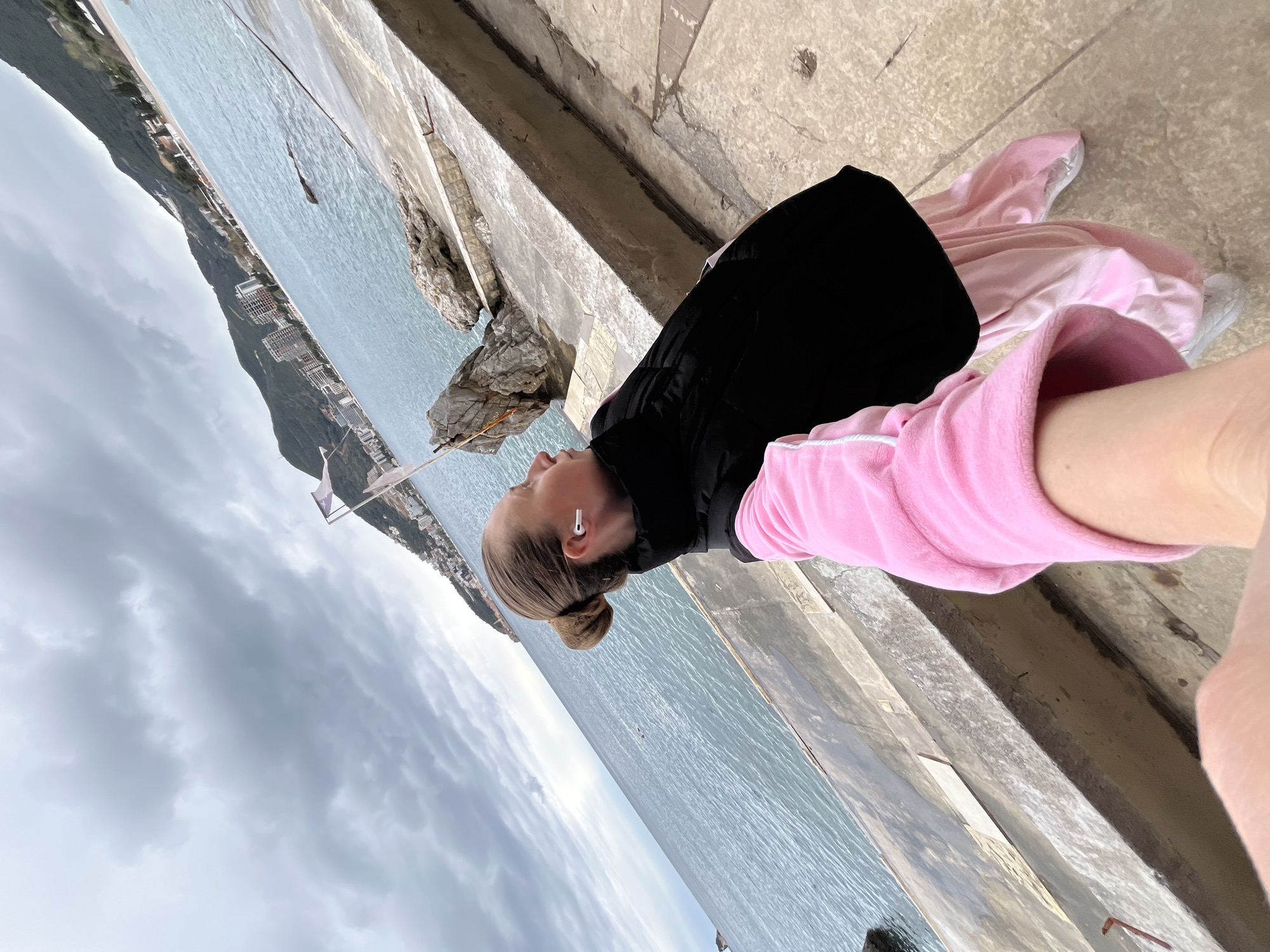 A woman in a black jacket and pink tracksuit lies on a concrete ledge by the sea. She has blonde hair pulled back in a bun and wears white earbuds. The water stretches to the horizon under a cloudy sky. In the distance, a coastline with buildings rises from the sea. This view is consistent with a location in Budva, Montenegro, given its coastal topography and building style. A concrete platform is to the woman's right.