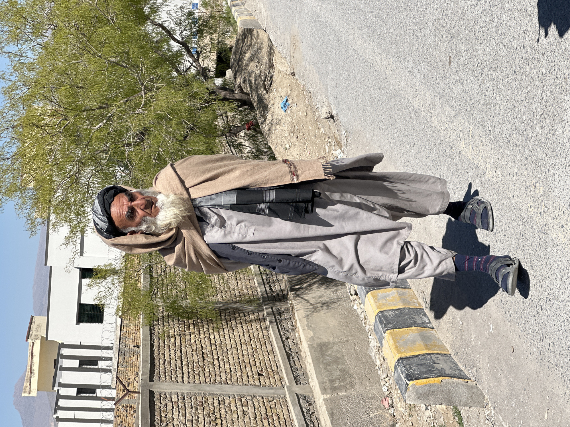 An elderly man stands on a paved road next to a yellow-and-black curb in Quetta City Tehsil, Pakistan. He wears a light gray shalwar kameez, a black and gray patterned shawl, and a black head covering. A light brown shawl is draped over his shoulders. The man has a long, white beard and dark skin. Behind him is a brick wall topped with barbed wire, with a white building visible beyond. A green tree stands to his left. The sky is pale blue. The ground is paved with asphalt.