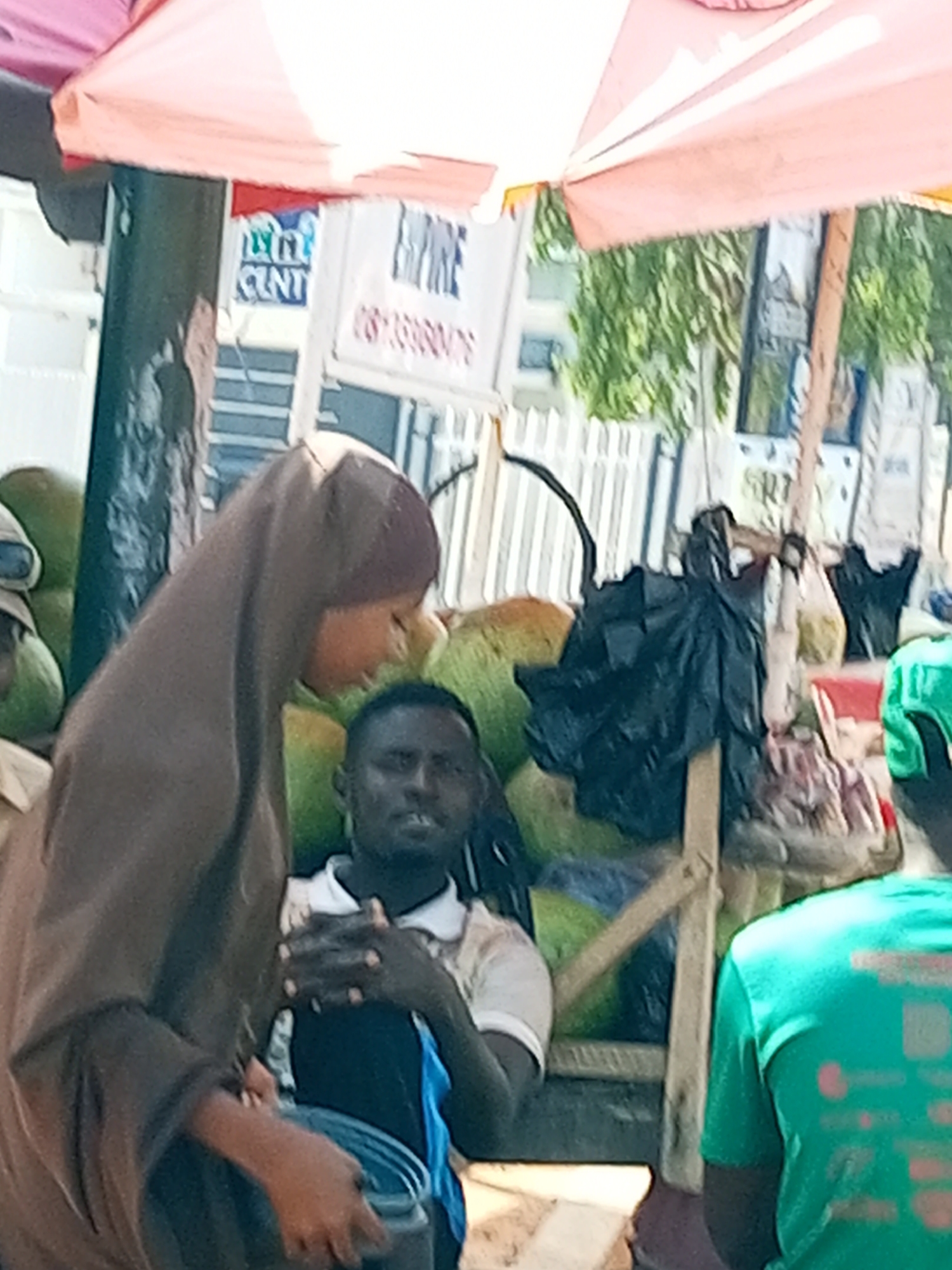 In Gombe, Nigeria, a scene unfolds under a pale pink umbrella, hinting at a market or street vendor setup. In the foreground, a woman wearing a brown hijab is partially visible, her face turned to the side, suggesting an interaction with someone or something out of frame. Her hands are engaged, holding a dark container, possibly a bucket, and reaching for its contents.
To her right, a man in a white polo shirt and a blue garment underneath is seated, his hands clasped together. His gaze is directed downwards, and his expression is difficult to discern. Behind him, a rough wooden stall supports an assortment of items, including large green fruits, resembling coconuts or gourds, and black plastic bags, perhaps containing goods or waste.
Further to the right, another individual is visible from the back, wearing a vibrant green t-shirt with an unclear graphic design. The environment appears to be outdoors, with hints of buildings and foliage in the background, indicating an urban setting. The lighting suggests it is daytime. A sign with some text, partially obscured by the umbrella, is visible behind the woman in the hijab, adding to the impression of a commercial area.