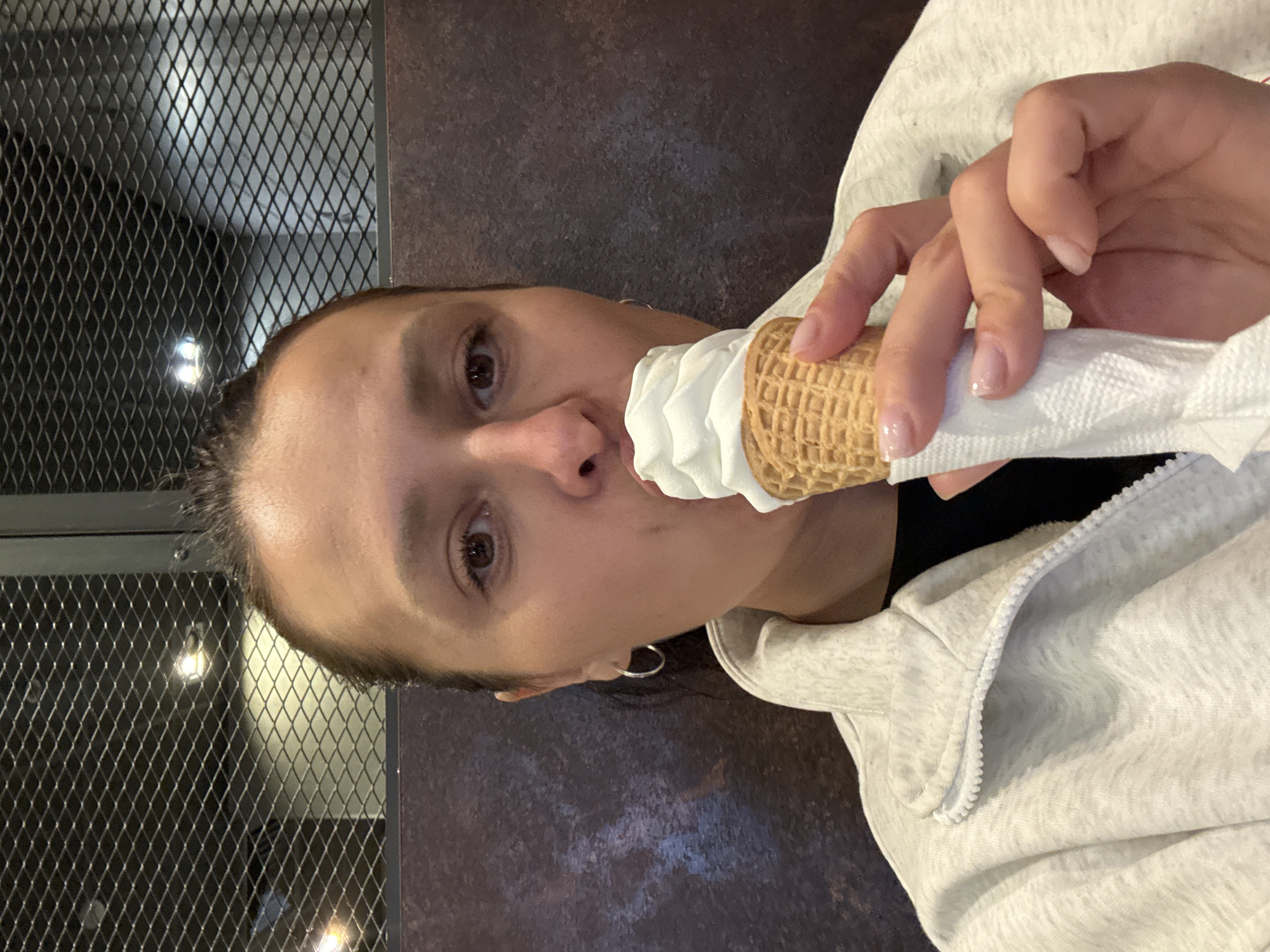A woman holds an ice cream cone to her mouth in an indoor setting. She wears a light grey zip-up hoodie and has hoop earrings. The background includes a wall with a metal grate and a darker, textured surface. The location is not discernible from the image and cannot be identified as Neftekamsk, Russia.
