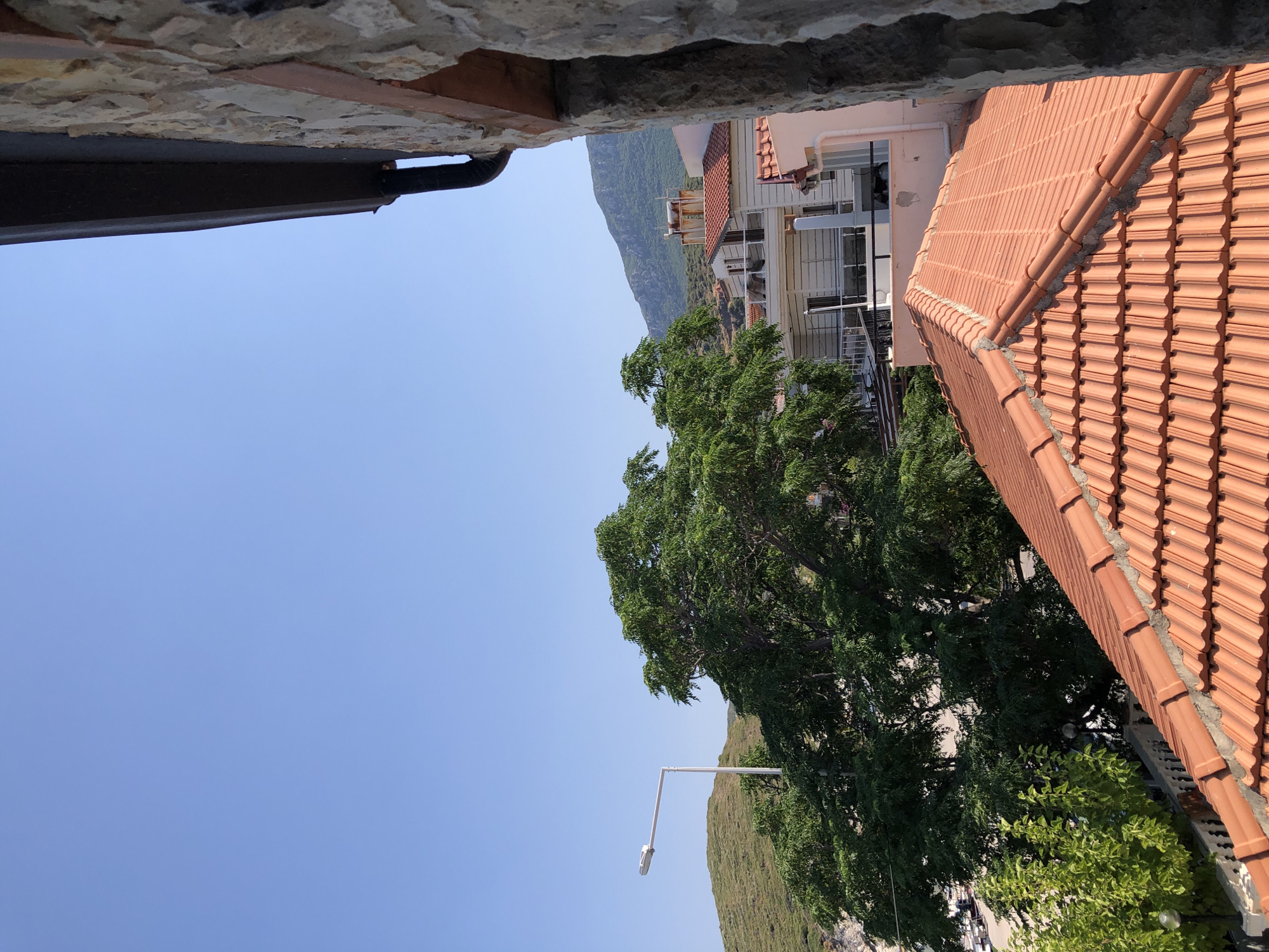 A street view in Karaburun, Turkey, features a clear blue sky above a hillside dotted with buildings and lush green trees. In the foreground, a terracotta tiled roof dominates the lower half of the image. To the left, a white streetlamp stands tall amidst vibrant green foliage, with a larger, mature tree to its right. Further in the background, several multi-story buildings with white facades and balconies are visible, their roofs also covered in red tiles. A majestic mountain range forms the backdrop, its slopes covered in dense green vegetation. The right side of the image is partially obscured by a stone wall and a dark gutter system. No people or animals are present in the scene.