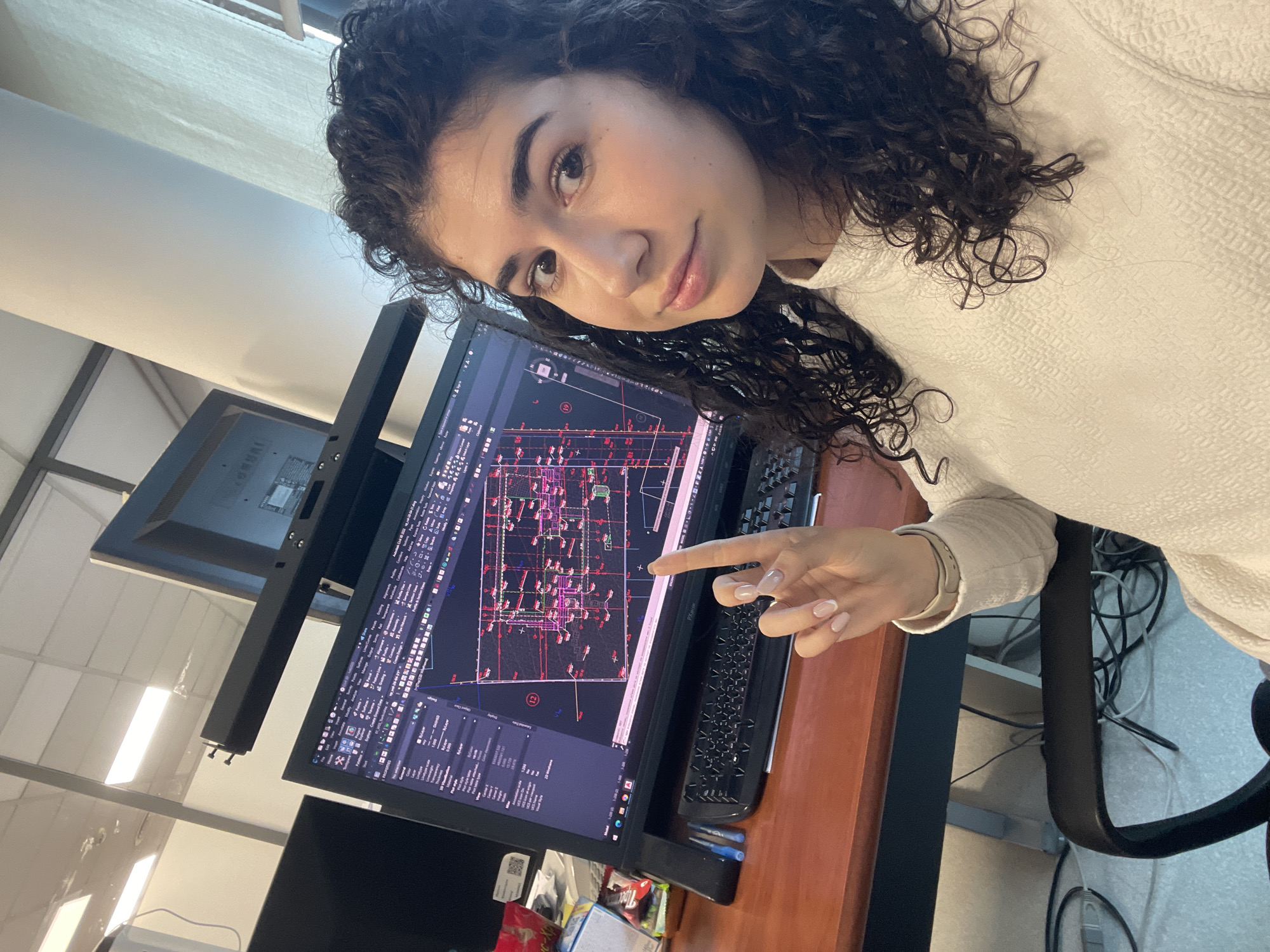 The image features a young woman in an office environment, captured at an angled perspective as if taken from a selfie. She has dark, curly hair and light brown eyes, looking towards the camera with a subtle, pleasant smile. She wears a cream-colored, textured long-sleeved top. Her right hand is visible, adorned with light pink, French-tipped fingernails, and she is pointing her index finger at a computer monitor.
The monitor displays a complex interface, which is identifiable as engineering or architectural CAD software, specifically "AutoCAD 2023," given the visible text. The screen shows detailed plans with lines, symbols, and numerical data in various colors (red, purple, white) against a dark background. Numerous software menus and tools are visible, including "File," "Edit," "View," "Insert," "Format," "Tools," "Draw," "Dimension," "Modify," "Parametric," "Window," and "Help." A computer keyboard is to the right of the monitor, and a second, partially visible monitor is positioned to the left.
The setting is an indoor office, characterized by bright, rectangular overhead lights reflected in the screens and on the ceiling. The woman is seated at a wooden desk, and the armrest of an office chair is visible in the foreground. Under the desk, a tangle of wires and cables can be seen. On the desk, near the base of the monitor, are partially visible items, including a blue box and a red and white plastic bag.
The woman's relaxed posture and subtle smile suggest a break or a posed moment during her work. The bright lighting indicates it is likely daytime. The overall scene depicts a professional workspace in Sofia, Bulgaria.