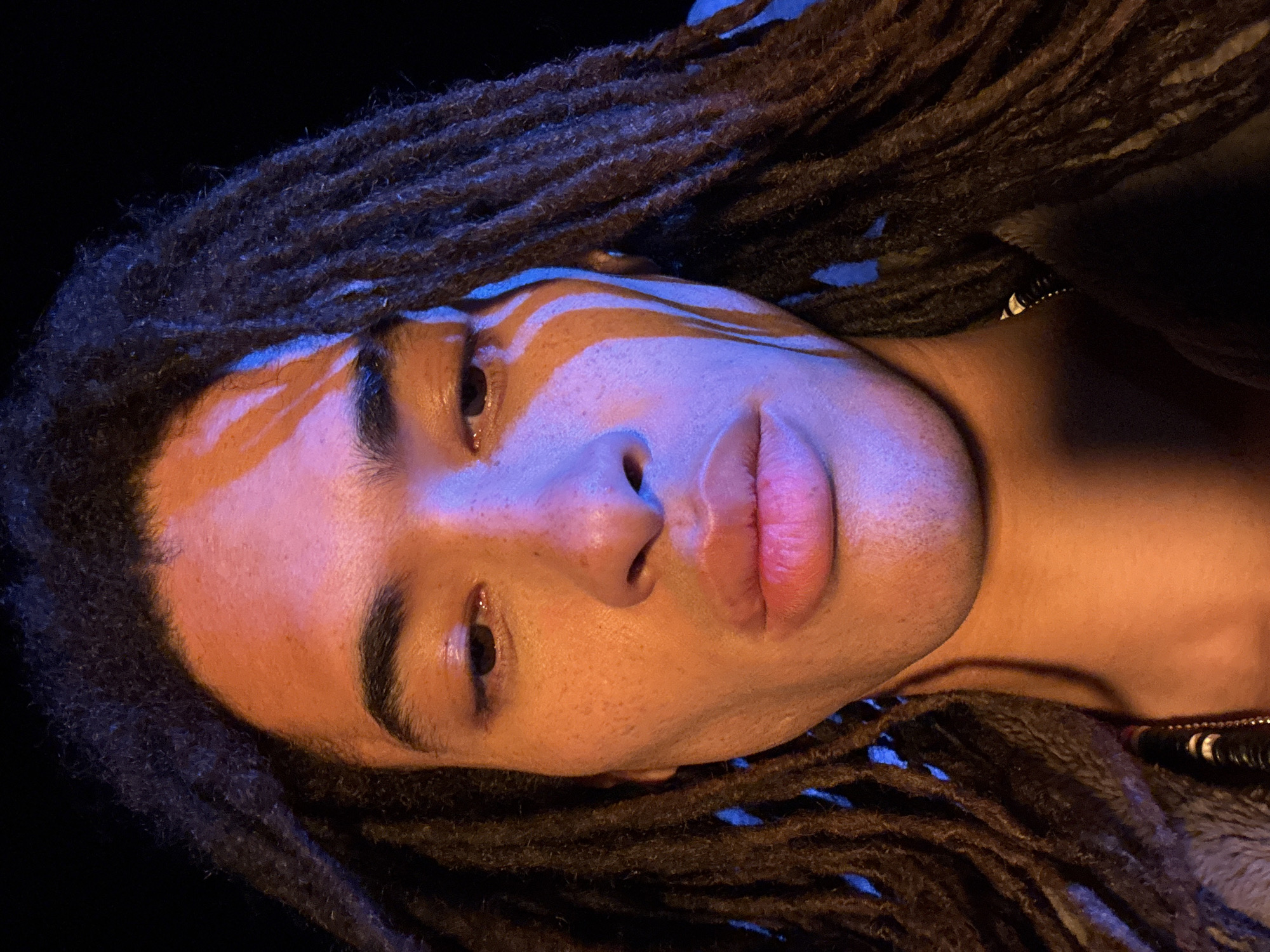A close-up, rotated headshot depicts a young person with dreadlocks, possibly in an urban environment. The individual has an olive complexion and full lips. A stark contrast of light and shadow falls across the face, with a possible pattern of light or paint visible above the eyebrow. The background appears dark and indistinct, suggesting nighttime or low-light conditions. The location cannot be reliably determined as Teguise, Spain based on the visual data alone.