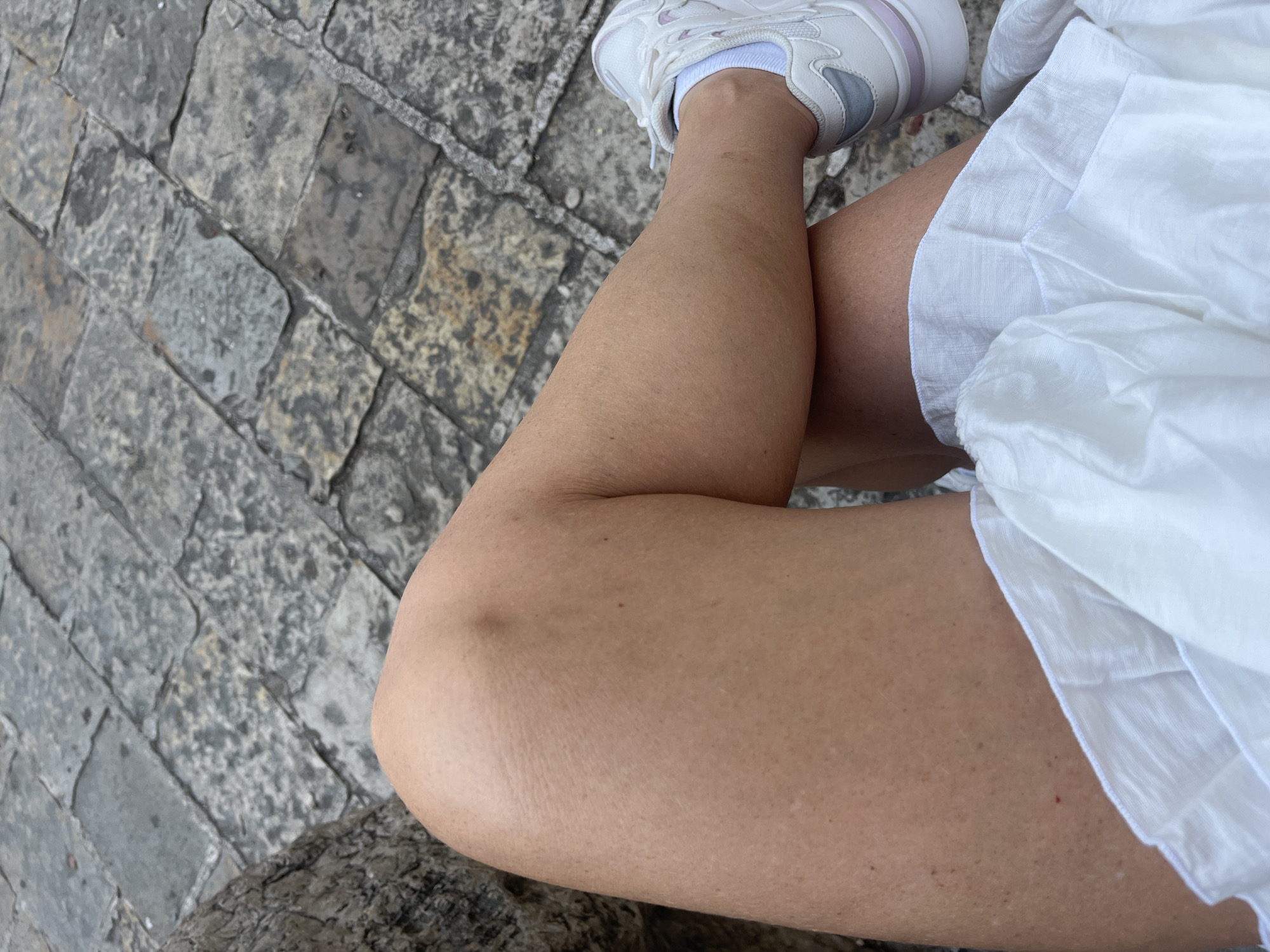 The image shows a close-up of a person's legs and white skirt, sitting on a stone pathway in Budva, Montenegro. The person is wearing white sneakers. The ground is paved with irregular grey stones. The focus is on the texture and color of the skin and the fabric of the skirt, with the stone pathway providing a textured background.