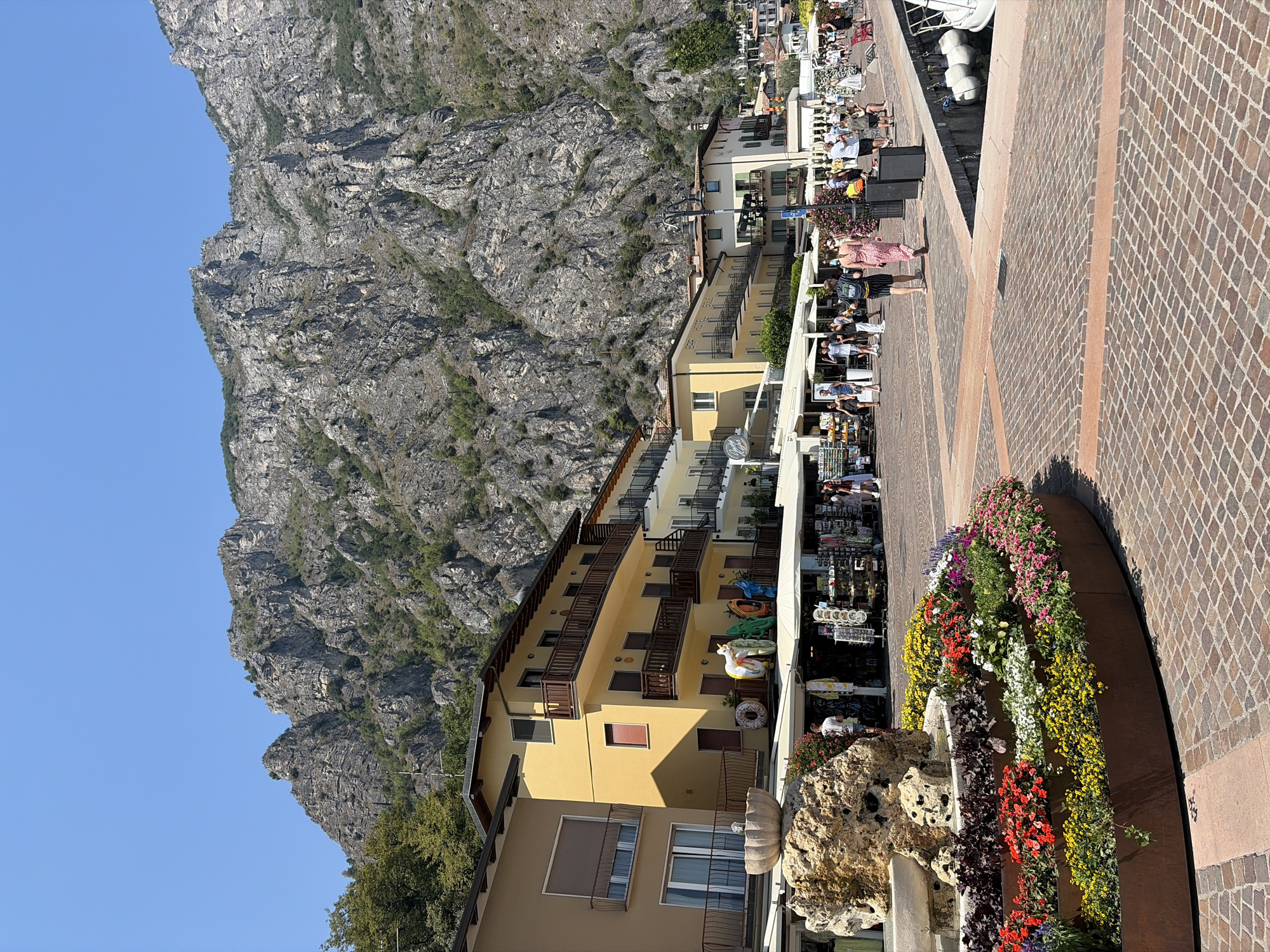 The image depicts a sunny day in the city of Limone sul Garda, Italy, likely on its lakefront promenade. In the foreground, a circular raised flower bed, filled with vibrant red, yellow, white, and pink flowers, sits on a cobblestone pavement. Behind the flower bed, a large rock formation with a small fountain is visible.
Adjacent to the promenade are several multi-story buildings with yellow and beige facades, featuring balconies with wooden railings. These buildings appear to house shops and possibly hotels, with awnings extending over storefronts displaying merchandise. A variety of people are strolling along the promenade, some browsing in the shops, others walking in pairs or groups. They are dressed in summer clothing.
In the background, a steep, rocky mountain range rises dramatically against a clear blue sky. The mountain is sparsely vegetated with trees and shrubs. The presence of boats docked along the waterfront, visible on the right side of the image, suggests the proximity of Lake Garda. The overall scene conveys a lively, tourist-oriented atmosphere in a picturesque lakeside town.