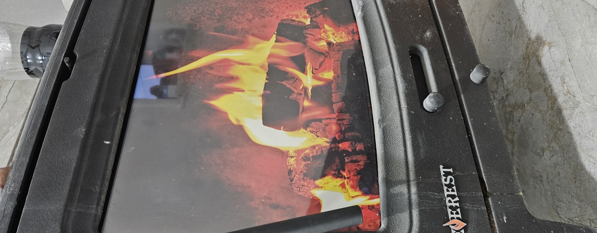 The image shows a close-up of a black electric fireplace with a realistic graphic of burning logs and flames displayed behind a glass screen. The fireplace has a textured surface and two dark, circular knobs on the lower section. The "EVEREST" logo is visible on the left side of the lower section. The fireplace is situated on a light-colored, textured floor, possibly concrete or stone, with some dirt and debris visible. In the upper right corner, a clear plastic bottle with a black lid, containing a dark liquid, is partially visible, resting on top of the fireplace. The background is dark and indistinct, with some faint reflections on the glass. The scene is indoors, and there are no people visible. This fireplace is likely located in a residential or commercial setting in Semey, Kazakhstan.