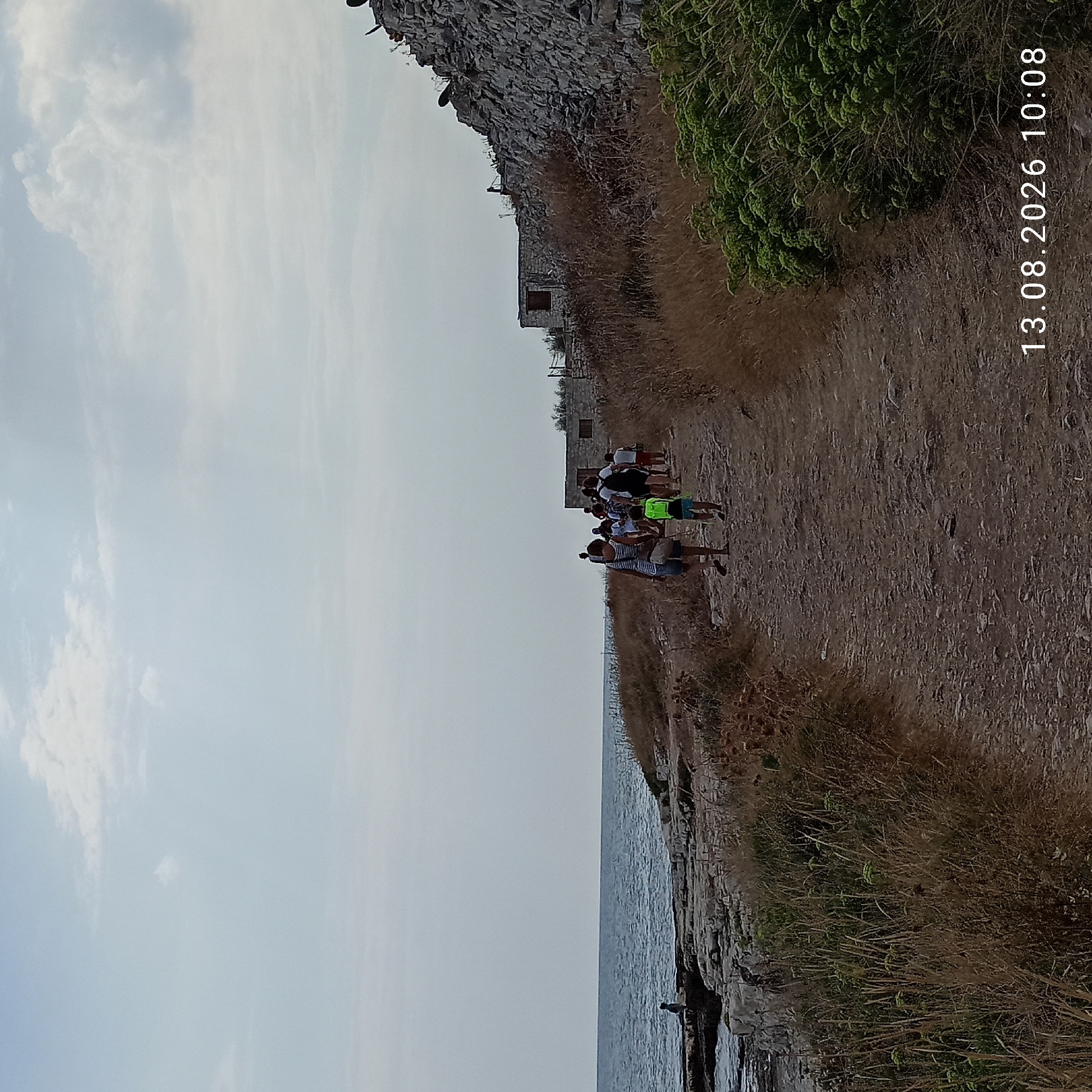 A group of people are walking along a dirt path on a rocky coastline. The path is lined with dry grass and some green bushes. In the background, there are stone buildings and a stone wall. The sea is to the left of the path, and the sky is overcast. The date stamp "13.08.2026 10:08" is visible in the lower right corner. This image depicts the area of the Old Port of Molfetta, Italy, likely near the remains of the ancient fortifications.