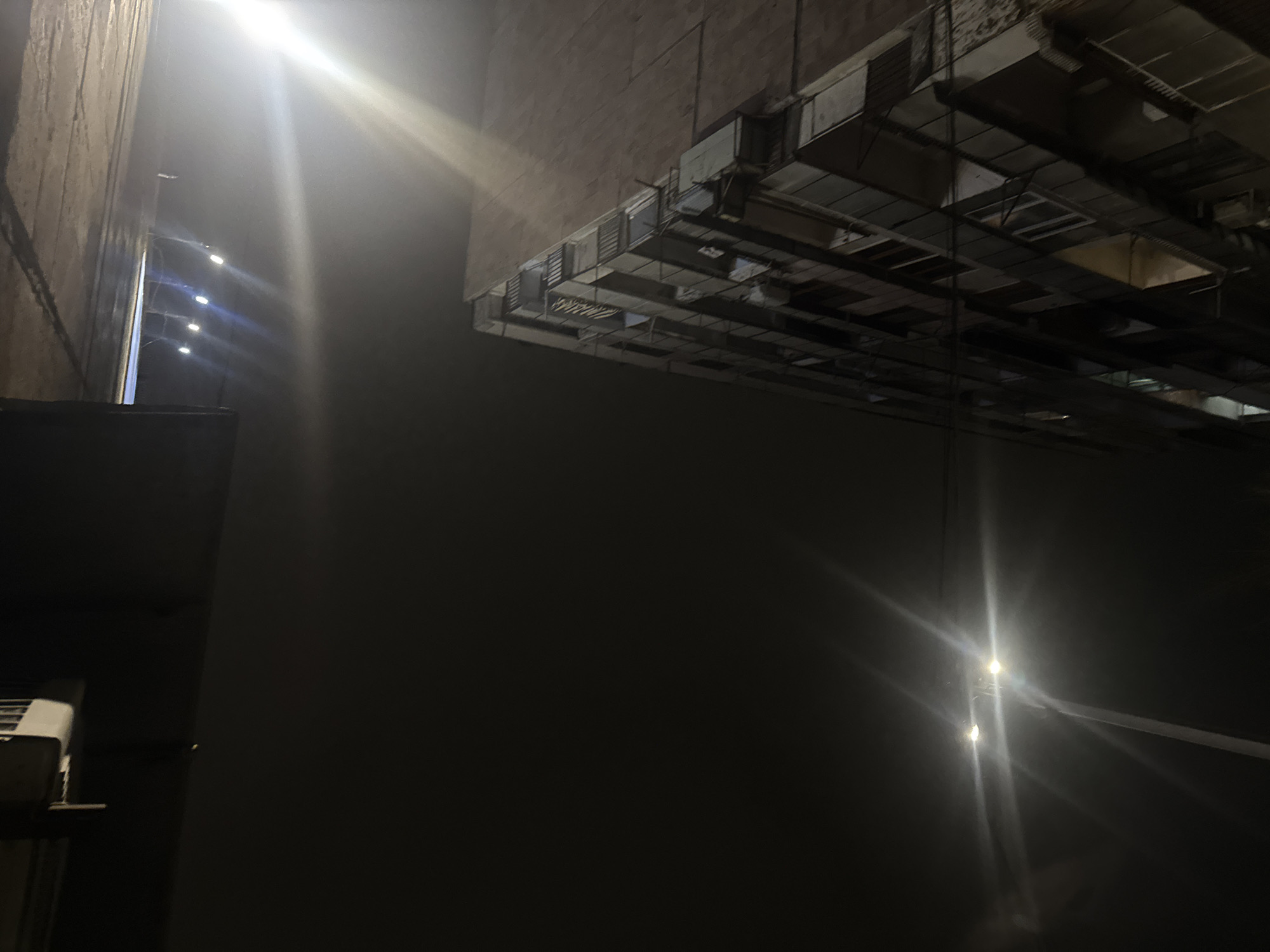 The image shows a night scene in what appears to be an industrial or commercial area. Overhead, a network of ductwork and metal supports creates a grid-like pattern against the dark sky. To the left, a tall, weathered building wall is visible, with what might be strings of lights or reflections along its surface. The lighting is harsh, with bright flares obscuring details and creating stark contrasts between light and shadow. The area is dark with faint illumination suggesting it is indoors or adjacent to a building. Due to the limited visual information available in the image, and without specific visual references to confirm, providing a precise location within Yerevan, Armenia, is not possible.