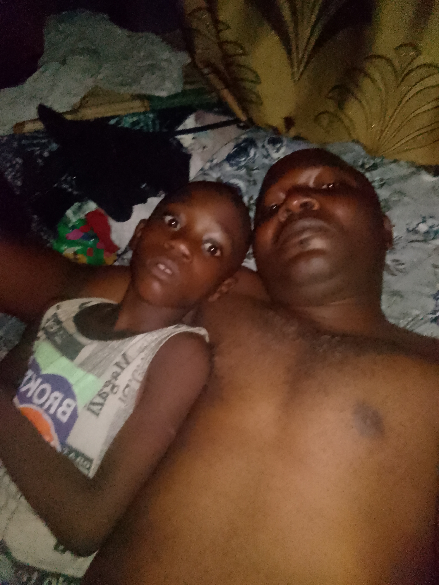 A man and a child are lying on a bed. The man is bare-chested and appears to be an adult. The child, who is wearing a tank top, lies next to him. The location is a bedroom, with bedding visible beneath them and clothing items and a curtain in the background. The specific city and broader location are unknown.