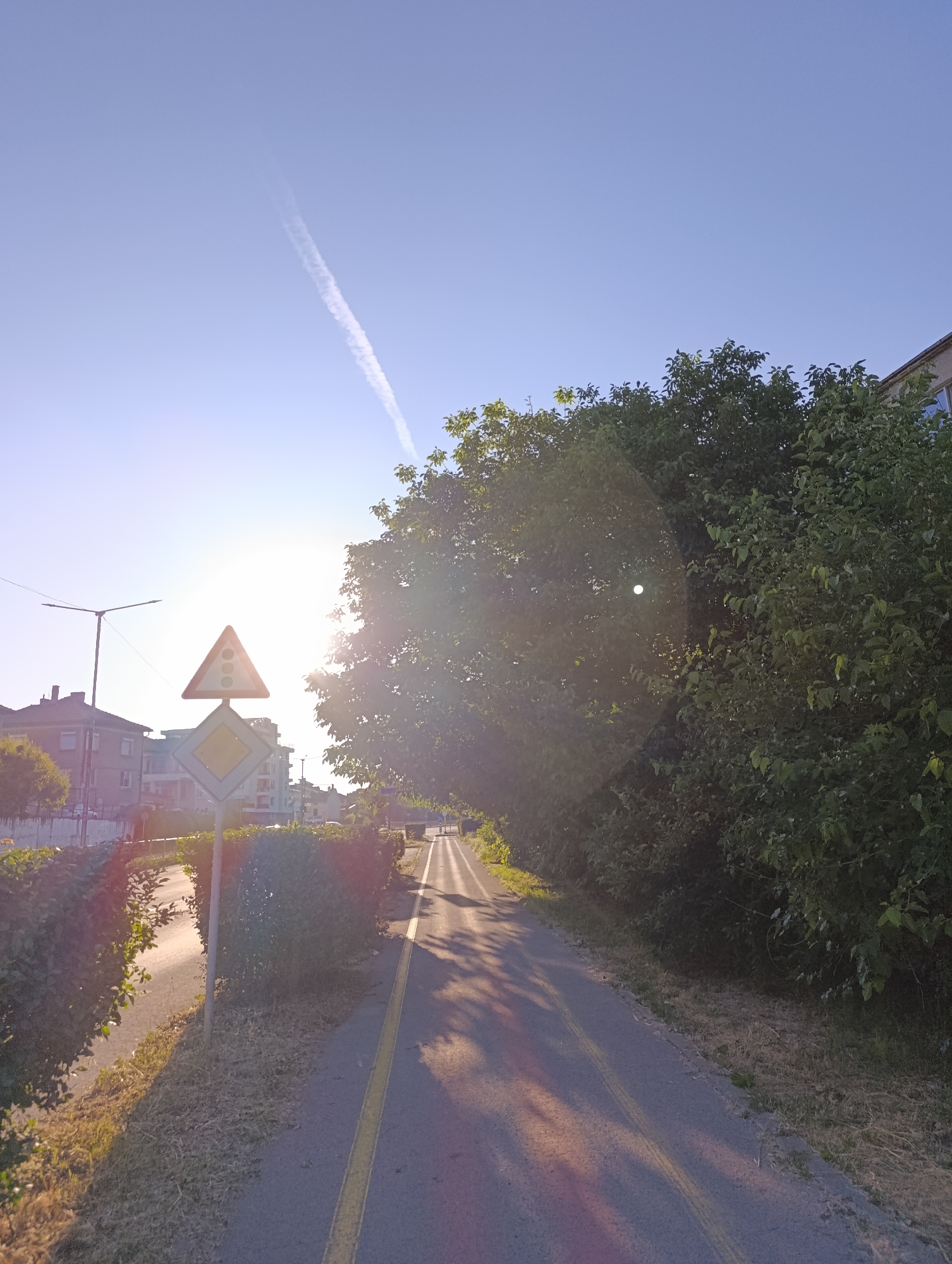 The image shows an outdoor scene with a clear blue sky, a contrail from an airplane, and the sun shining brightly. In the foreground, there is a paved path with yellow lines, suggesting it could be a bike path or a pedestrian walkway. To the left of the path, there are traffic signs, including a triangular sign with three circles (likely indicating traffic lights) and a diamond-shaped sign below it. Adjacent to the path, there are hedges and trees, with lush green foliage. In the background, buildings can be seen, with some appearing to be residential homes. The sunlight creates lens flare and casts long shadows across the path, indicating that it is either early morning or late afternoon. The overall mood is peaceful and serene, with the bright sunshine and clear sky suggesting fair weather. There is no visible text in the image.